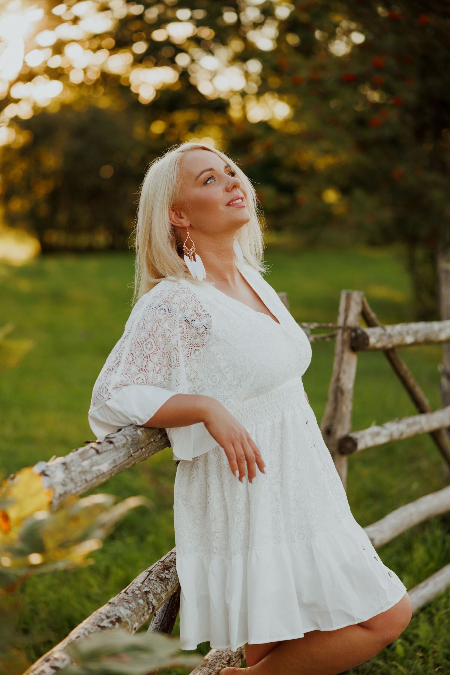 Woman in white lace dress poses against rustic fence at golden hour.
