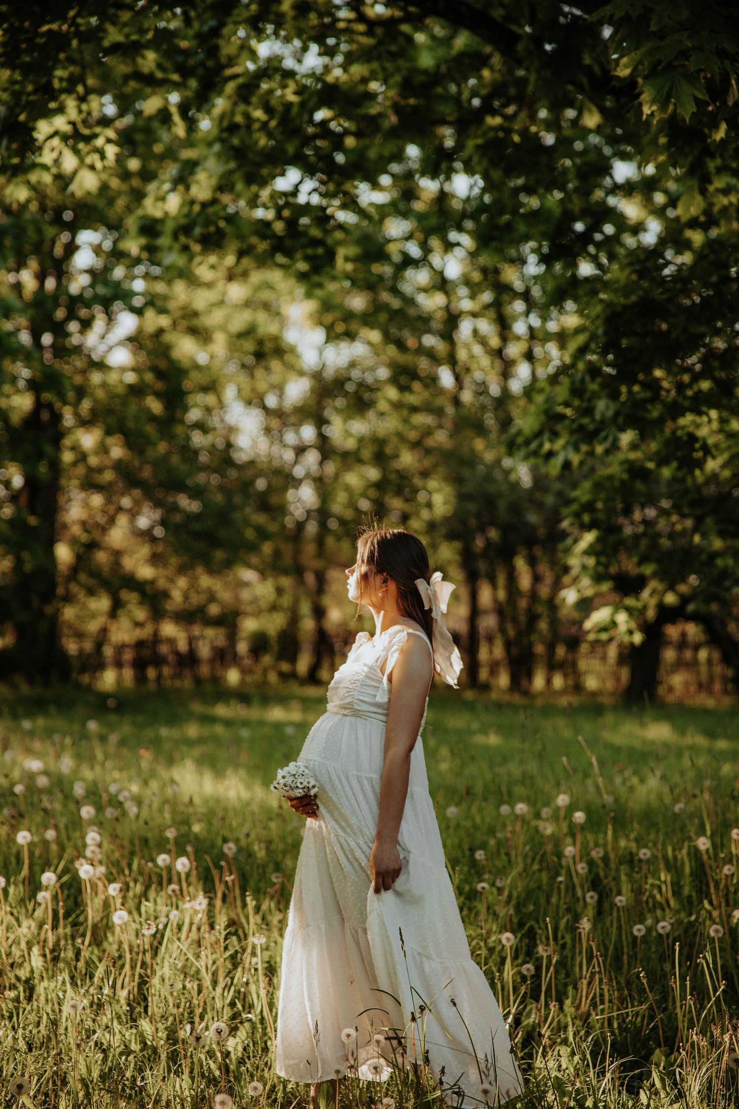Ethereal maternity portrait in flowing white dress amid sunlit meadow.