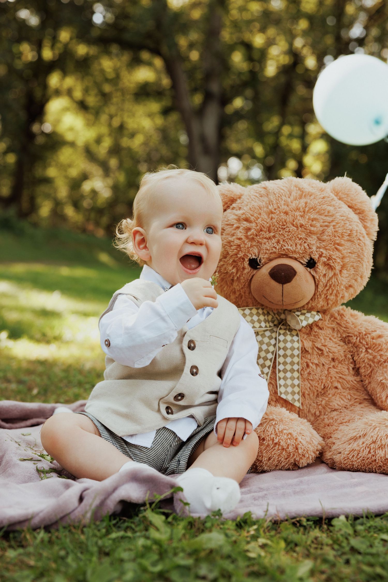 Baby lying on blanket outdoors with large teddy bear toy showing joyful expressions.