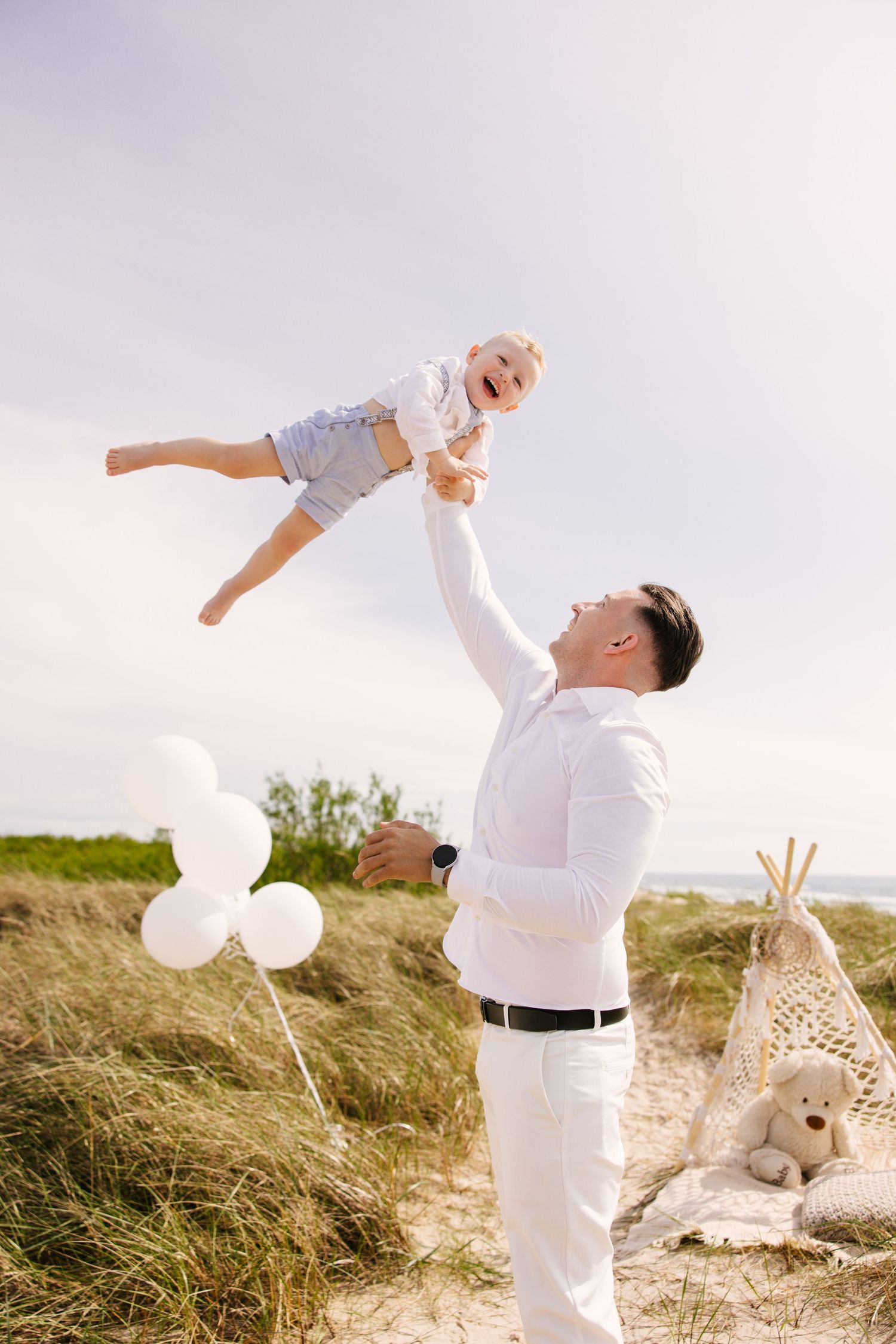 Parent and child in white clothing playing together with balloons on a grassy beach dune during sunset.
