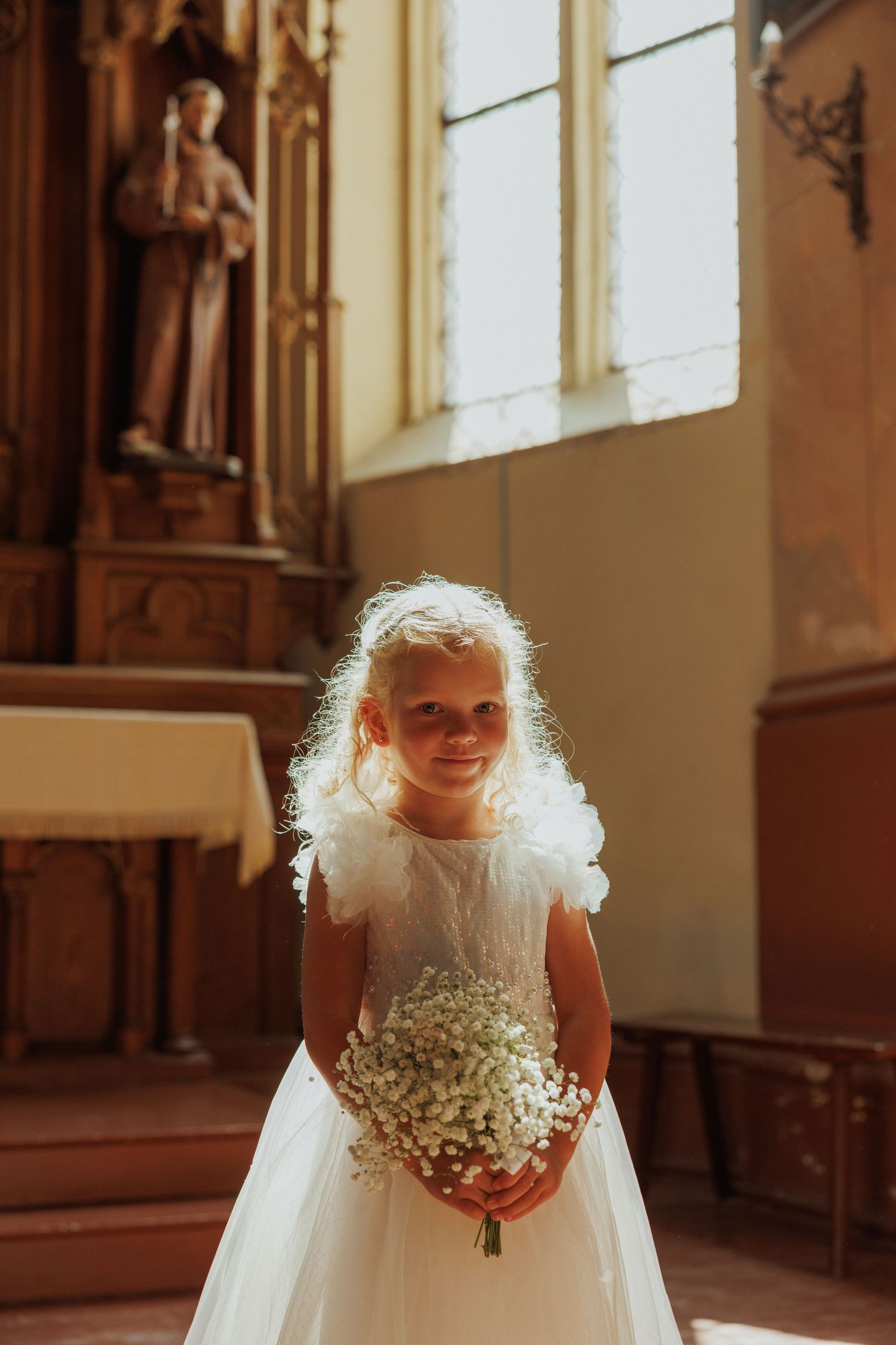 Young attendant in white dress holding baby's breath flowers in an ornate church setting.