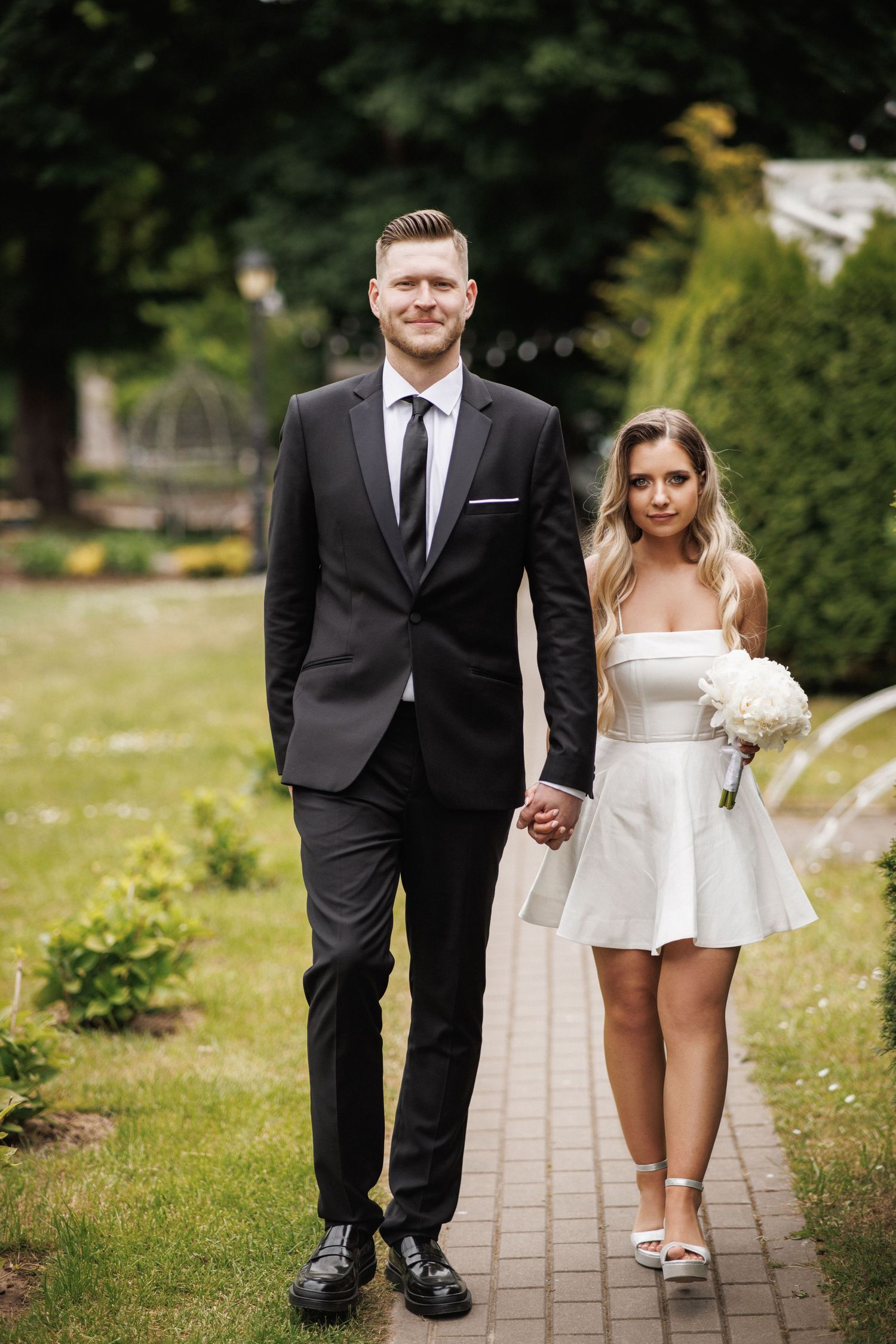 A couple in formal wedding attire poses together on a stone path surrounded by green grass and trees.
