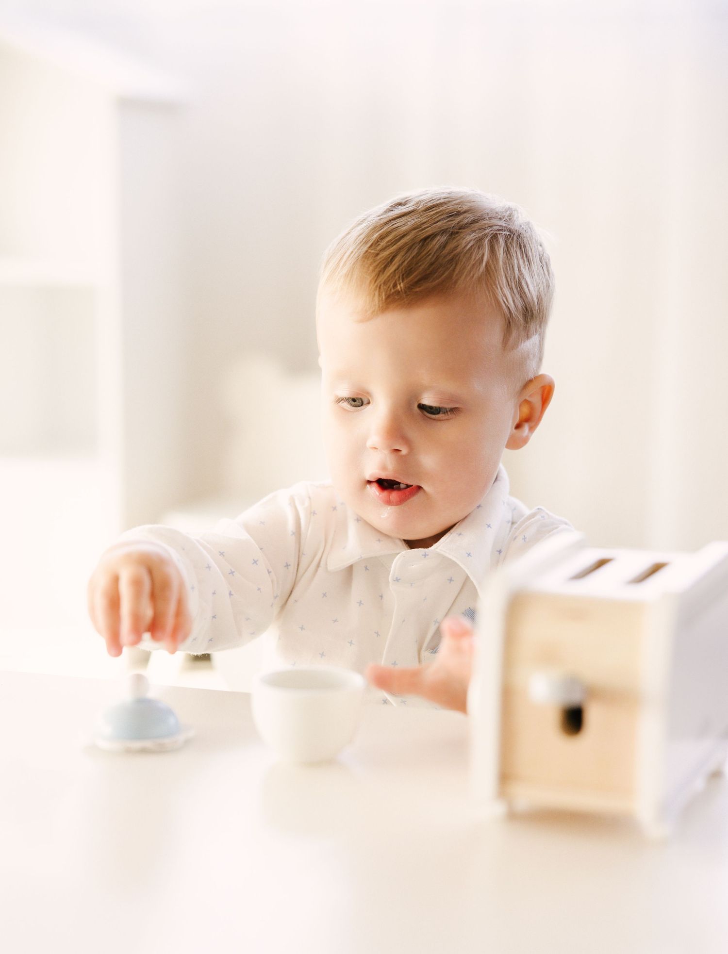 Small child in white shirt reaching for objects on a bright white table with natural lighting.