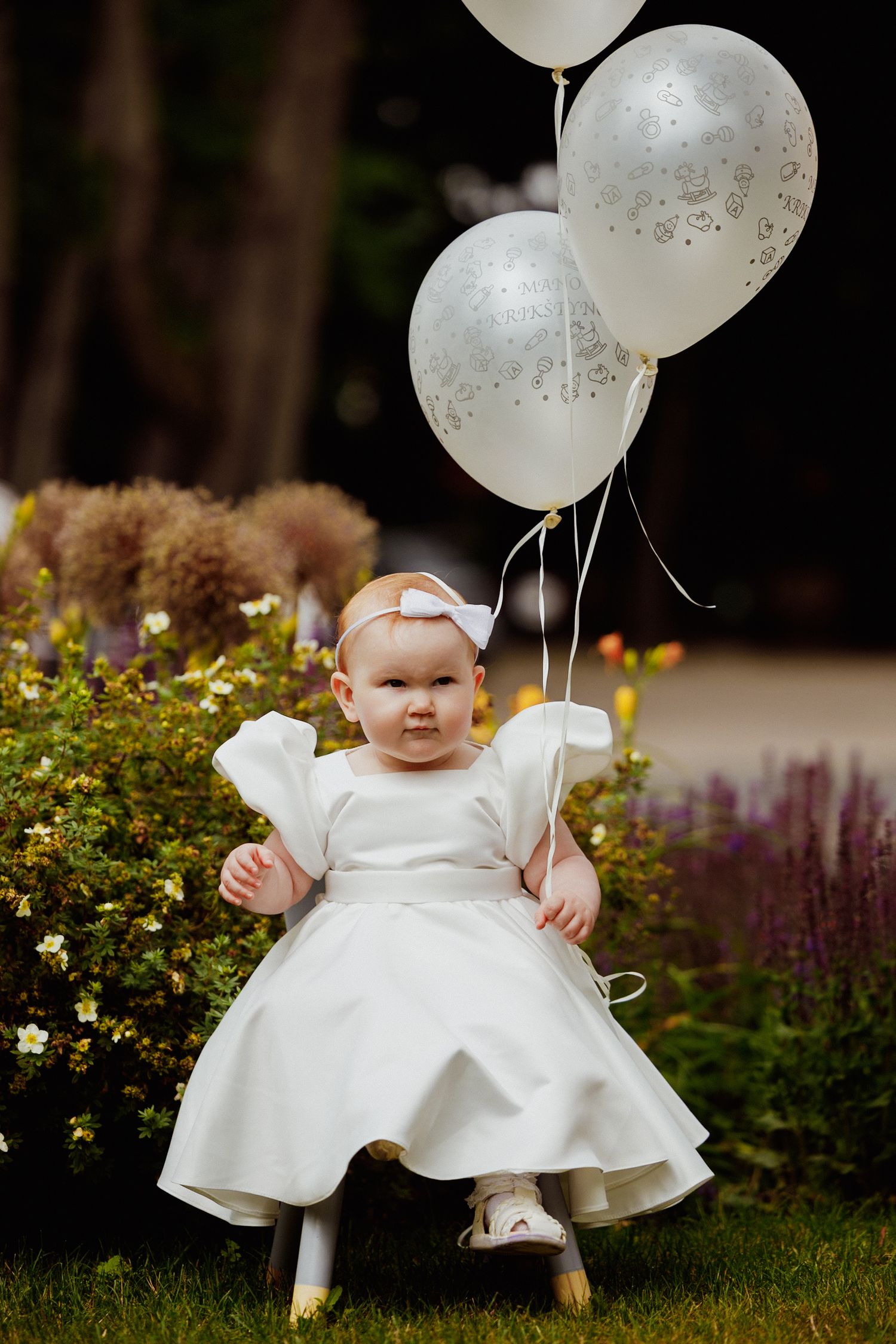 Small child in white christening dress holding two white balloons stands in garden with blooming flowers.