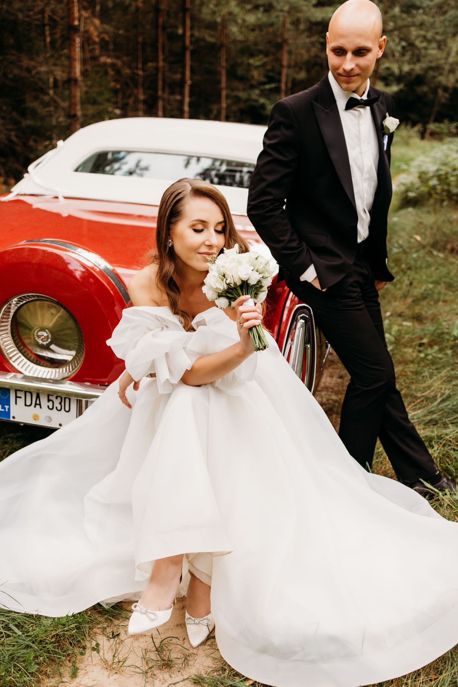 A romantic wedding photo shoot featuring a flowing white dress and a classic red vintage car in a forest setting.