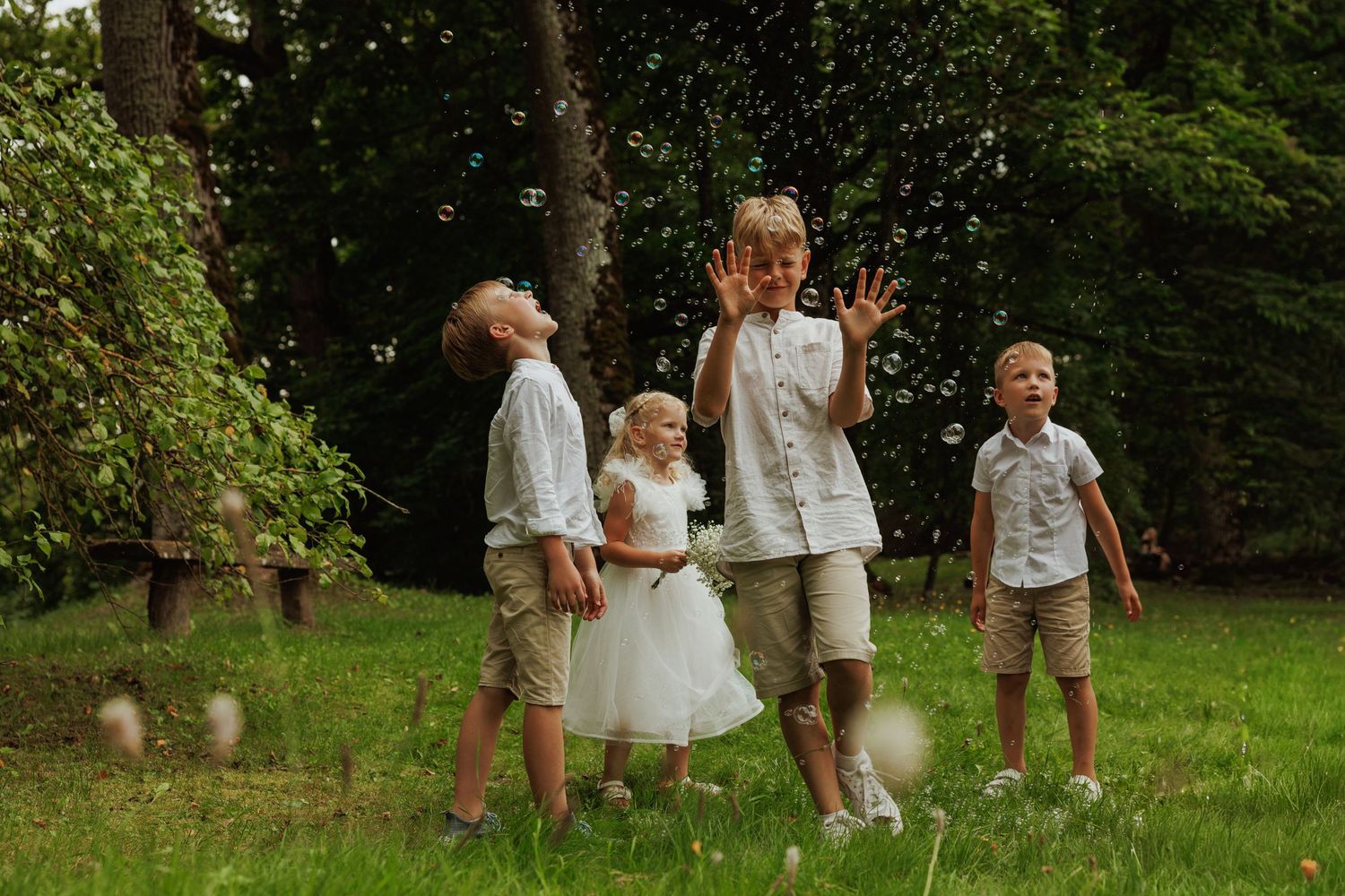 Children in khaki and white outfits play and run together in a lush green garden setting during a summer celebration.
