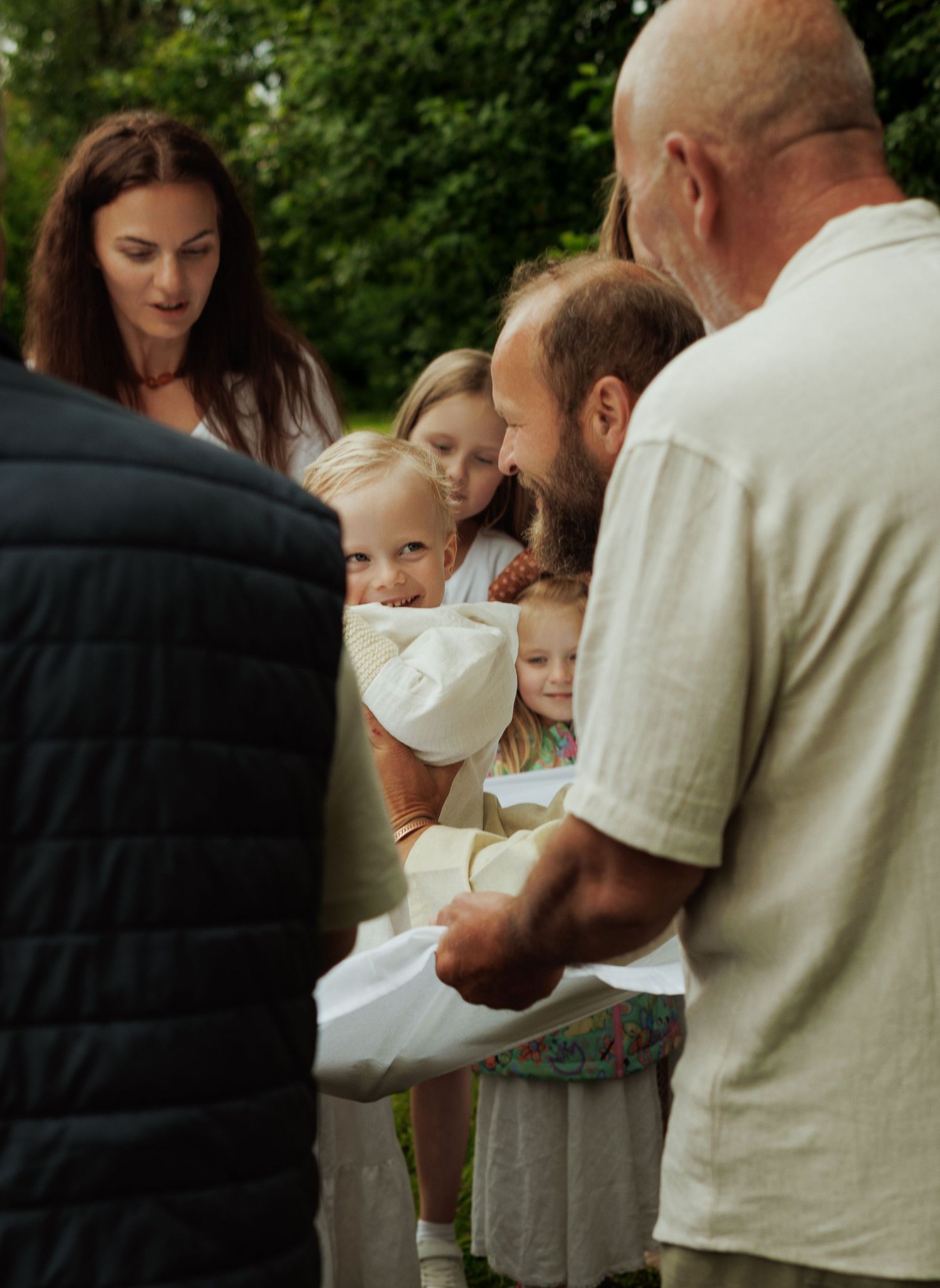 Family gathering outdoors with adults and children in white attire, sharing a warm moment in a lush green setting.