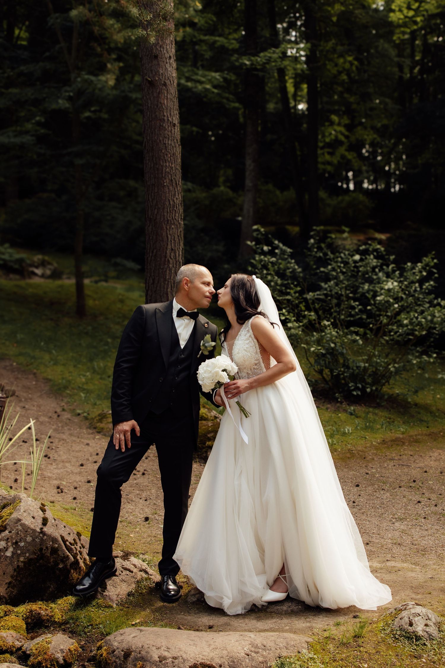 A bride and groom share a romantic kiss in a lush forest setting, she in a white gown, he in a black tuxedo.