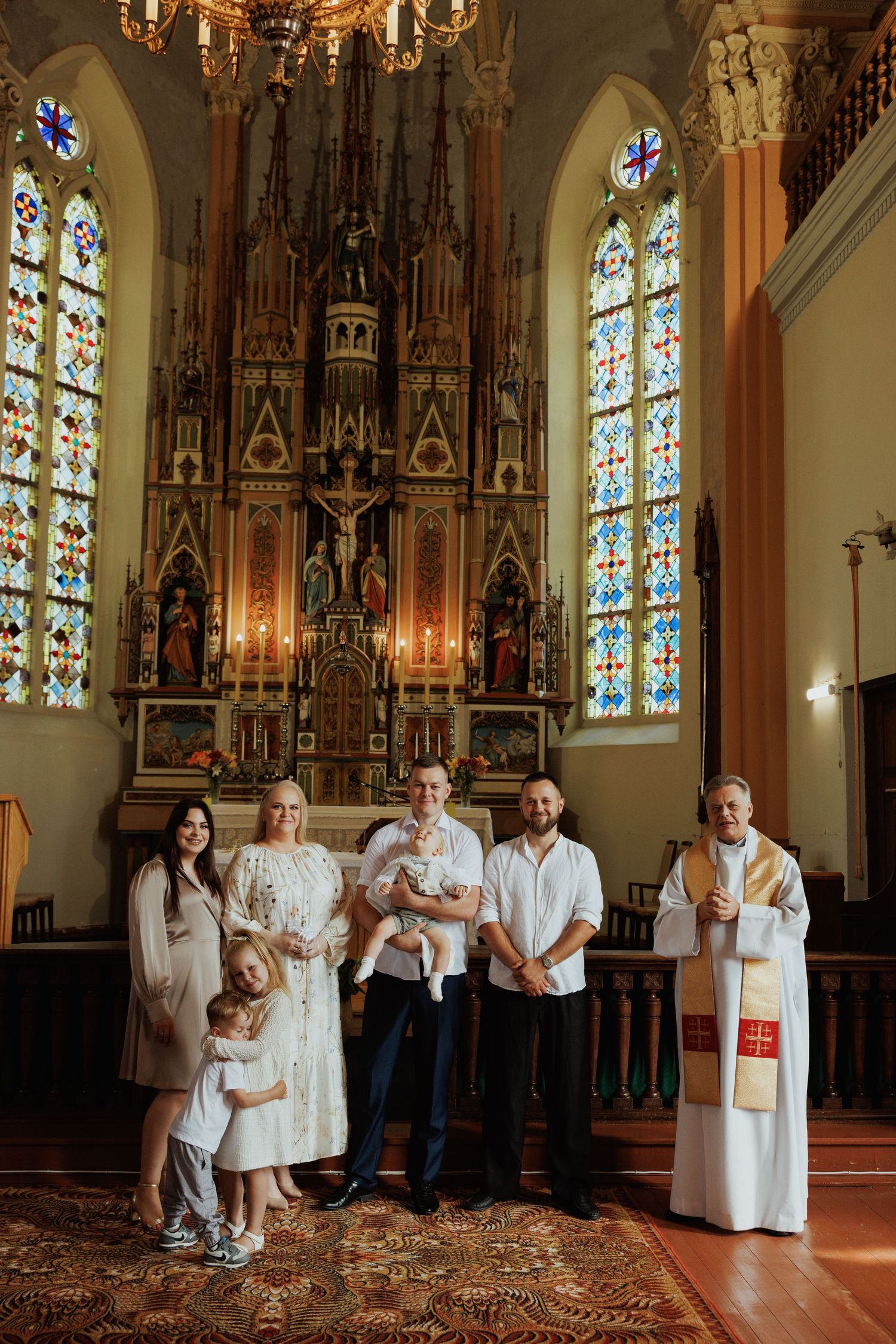 A baptism ceremony takes place in a beautiful Gothic church with stained glass windows and an ornate altar.