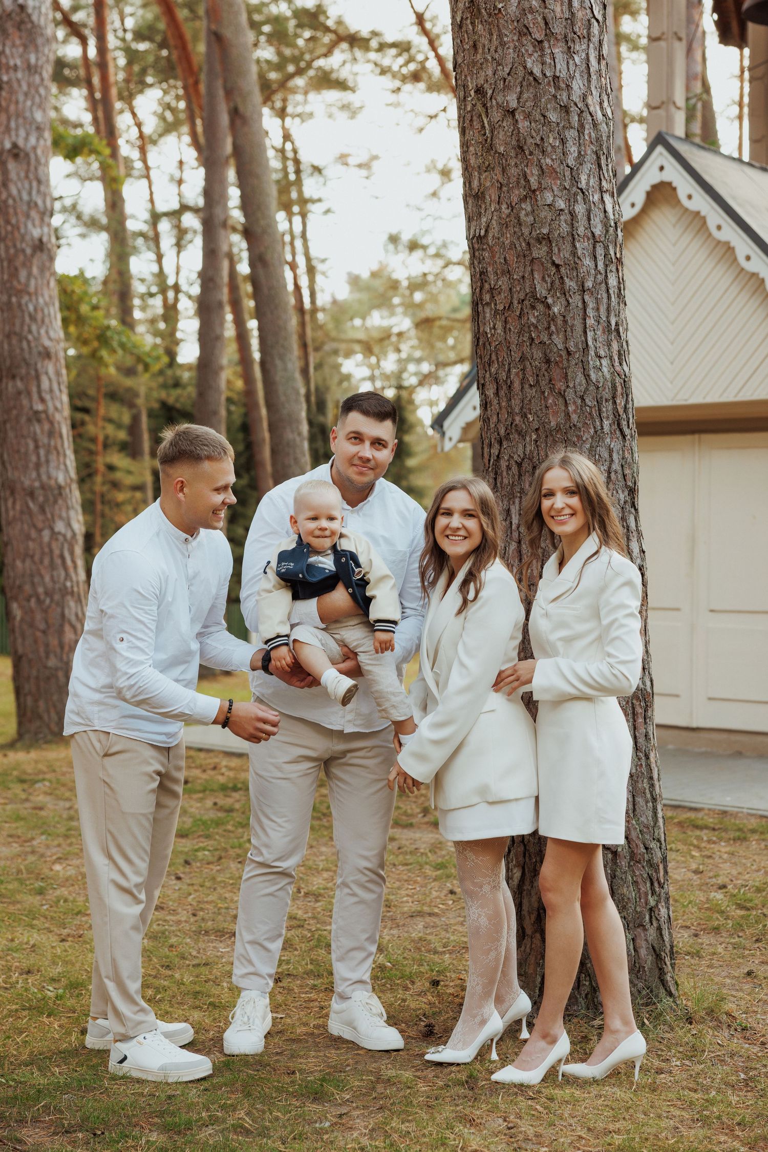 Family wearing coordinated white outfits poses together in a pine forest setting for outdoor photo session.