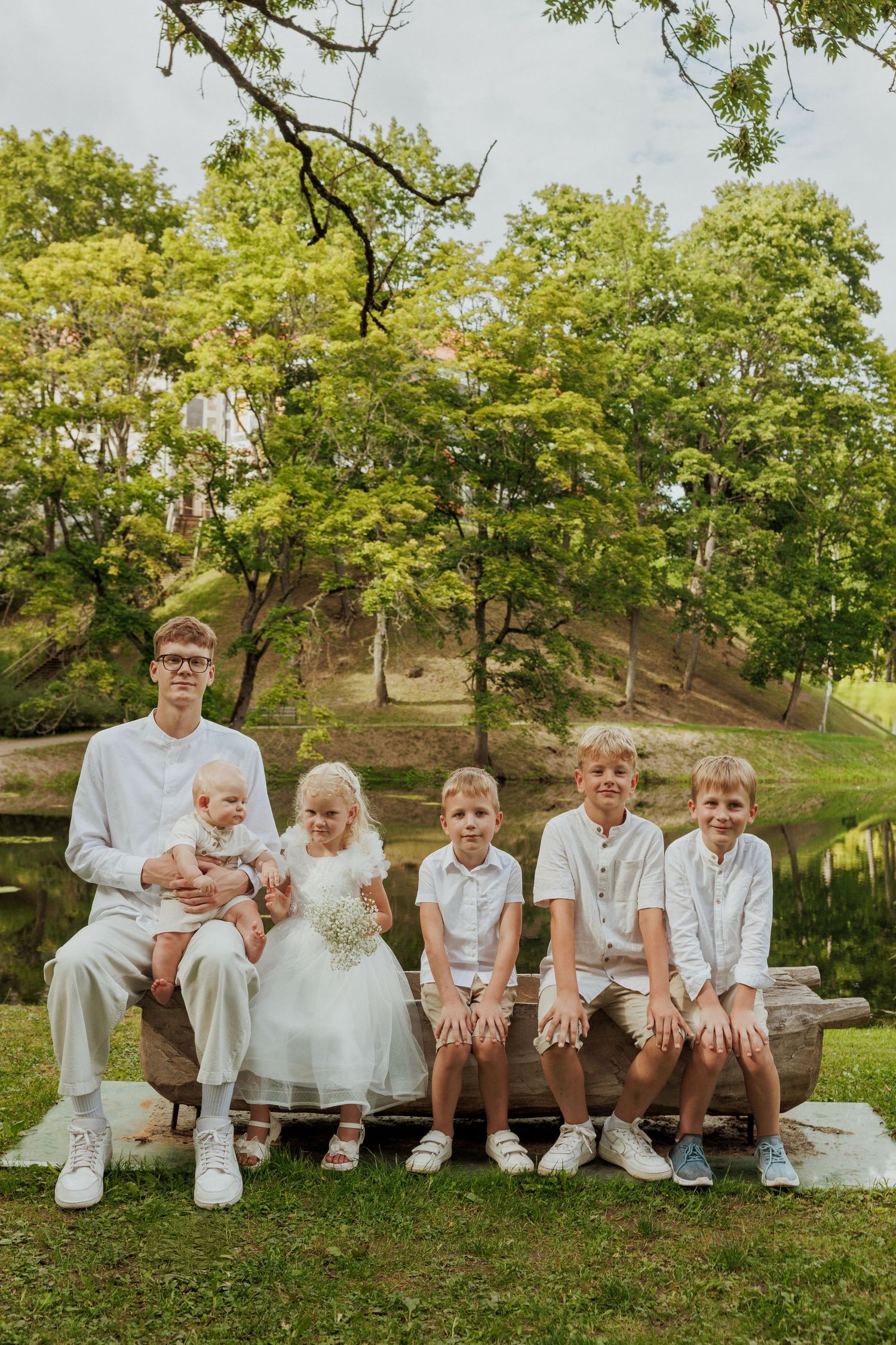 A group in coordinated white attire sits together on a wooden bench in a lush outdoor setting.