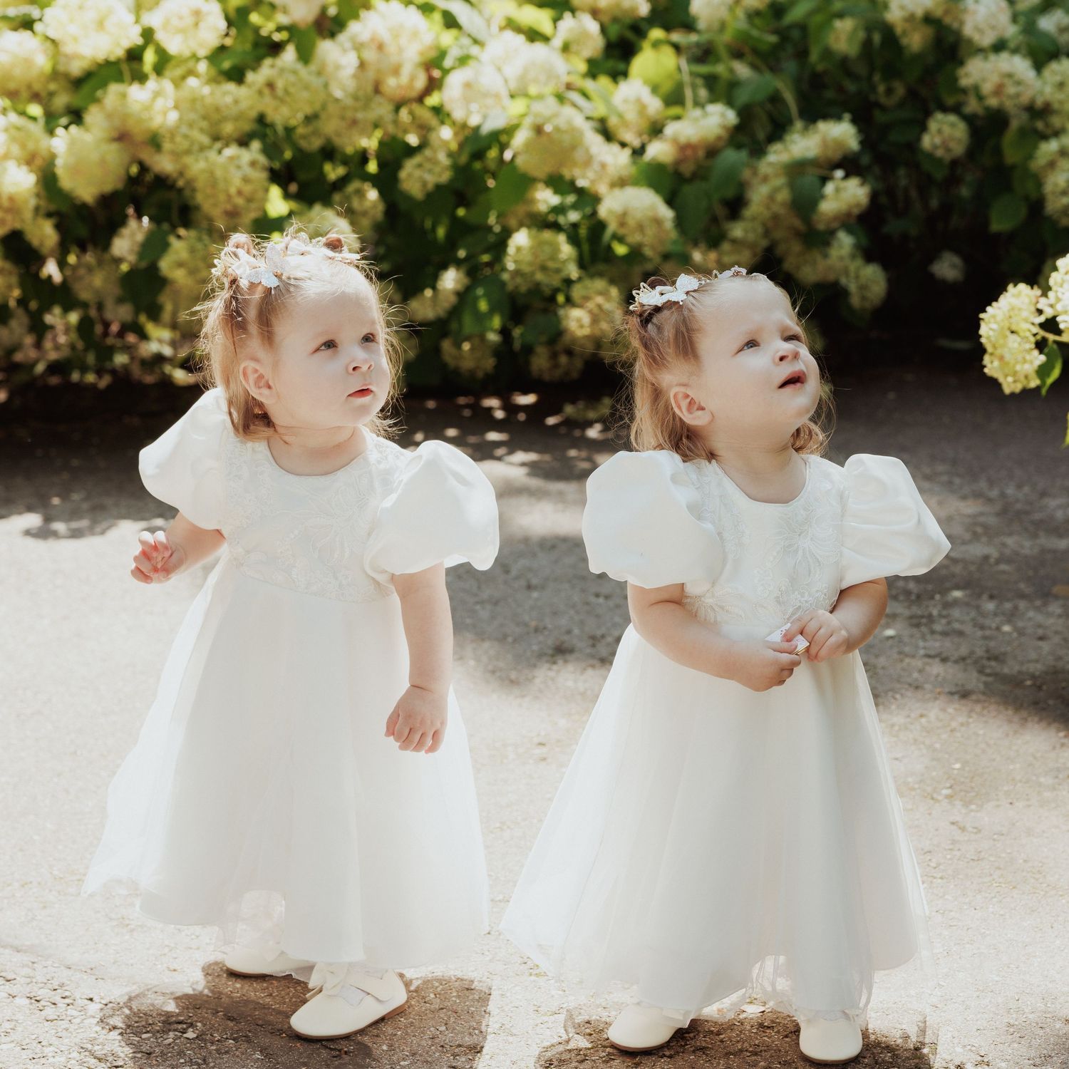 Two small children in matching white dresses and shoes stand together near blooming white flowers in a garden setting.