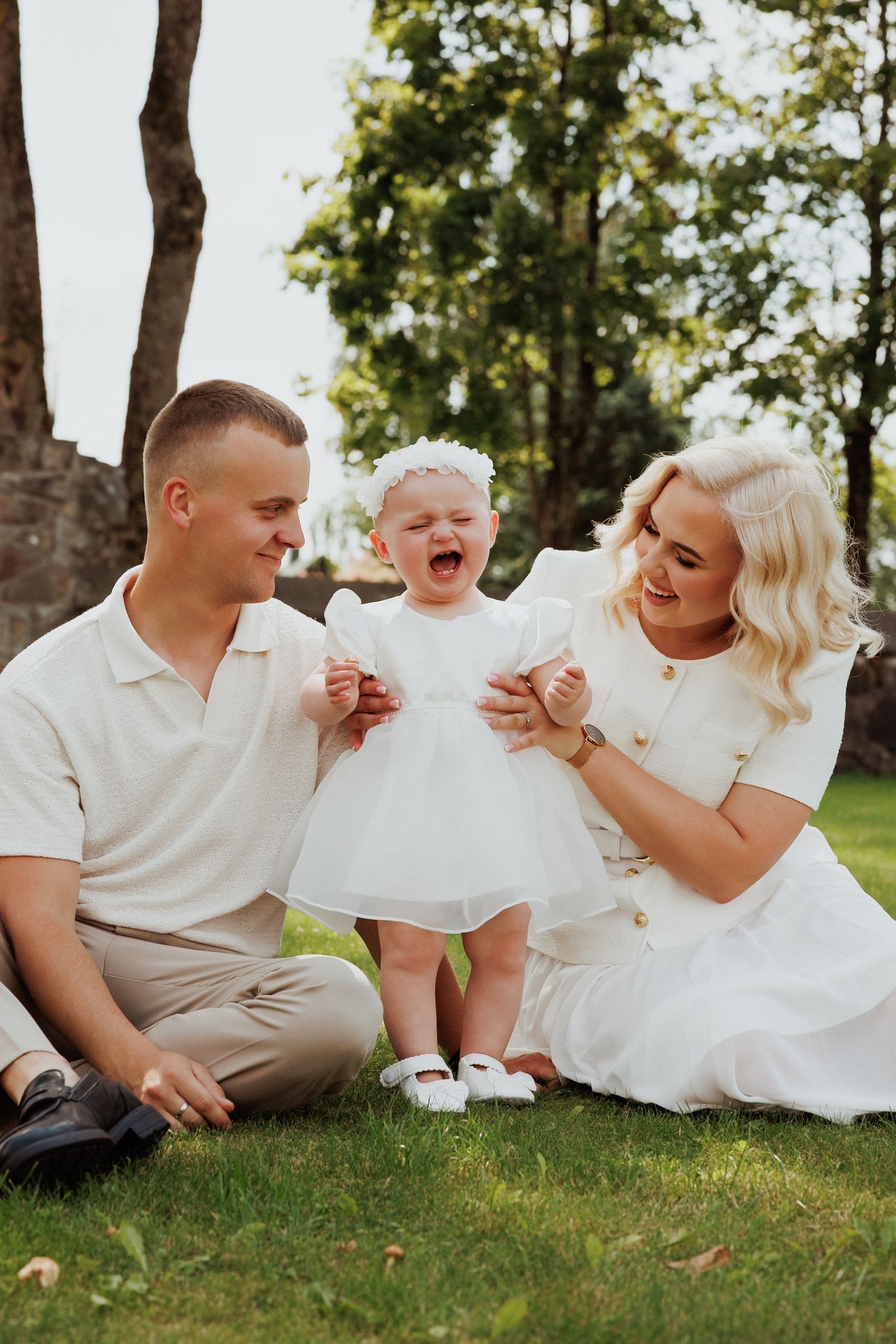 Family dressed in white shares playful moments while sitting on grass under trees.
