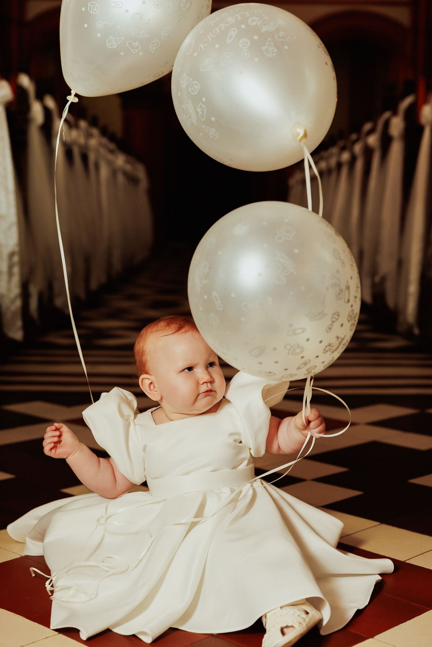 Baby in white dress lying on checkered floor with white balloons floating above.