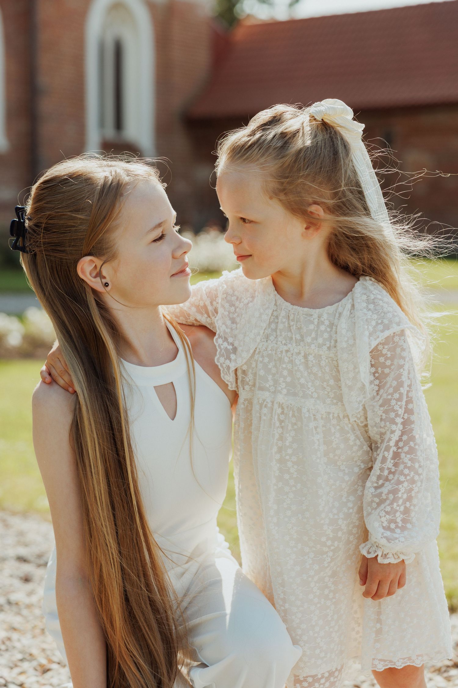 Two young girls in white dresses share a sweet moment together outdoors.