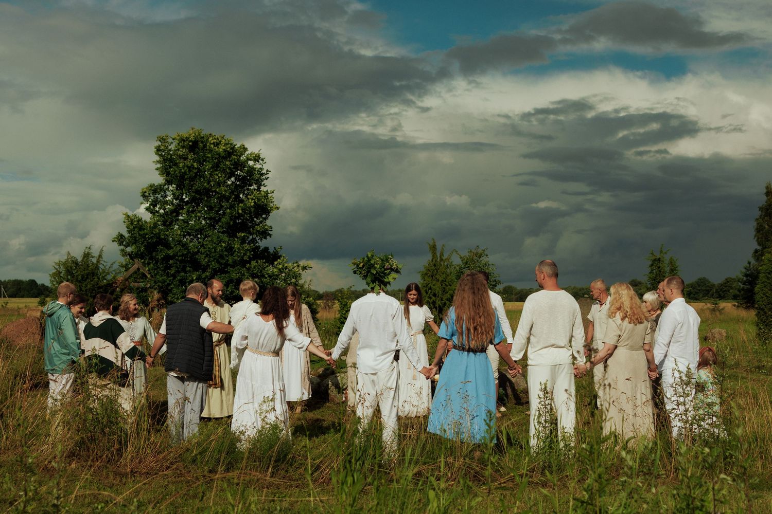 Group of people holding hands in a circle outdoors in a green field under dramatic cloudy sky wearing white attire.