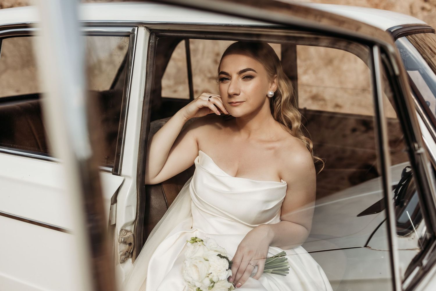 A bride in a white wedding dress sits elegantly in a vintage car holding a bouquet of white flowers.