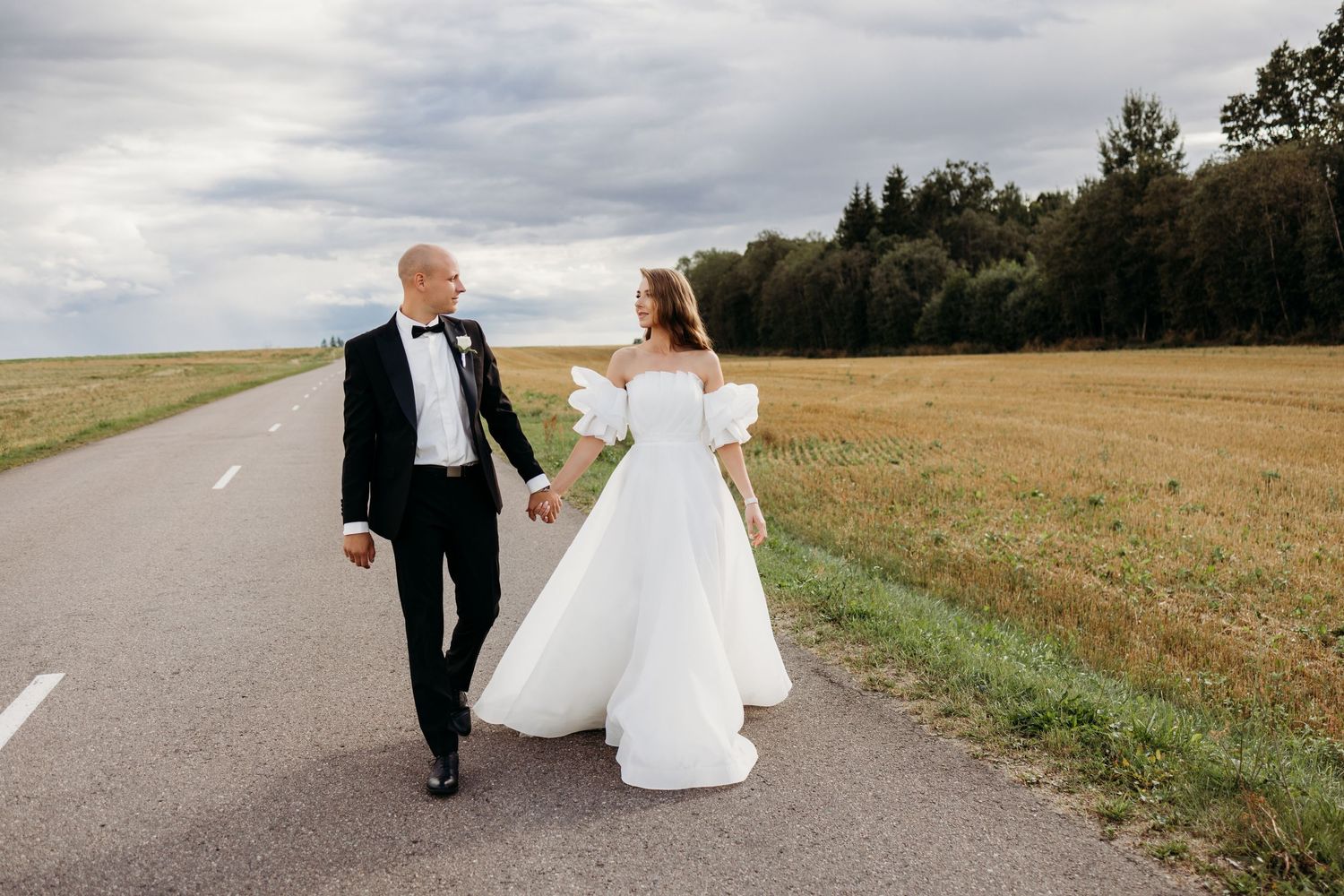 A couple walks hand in hand along a rural road with fields and cloudy skies in the background.