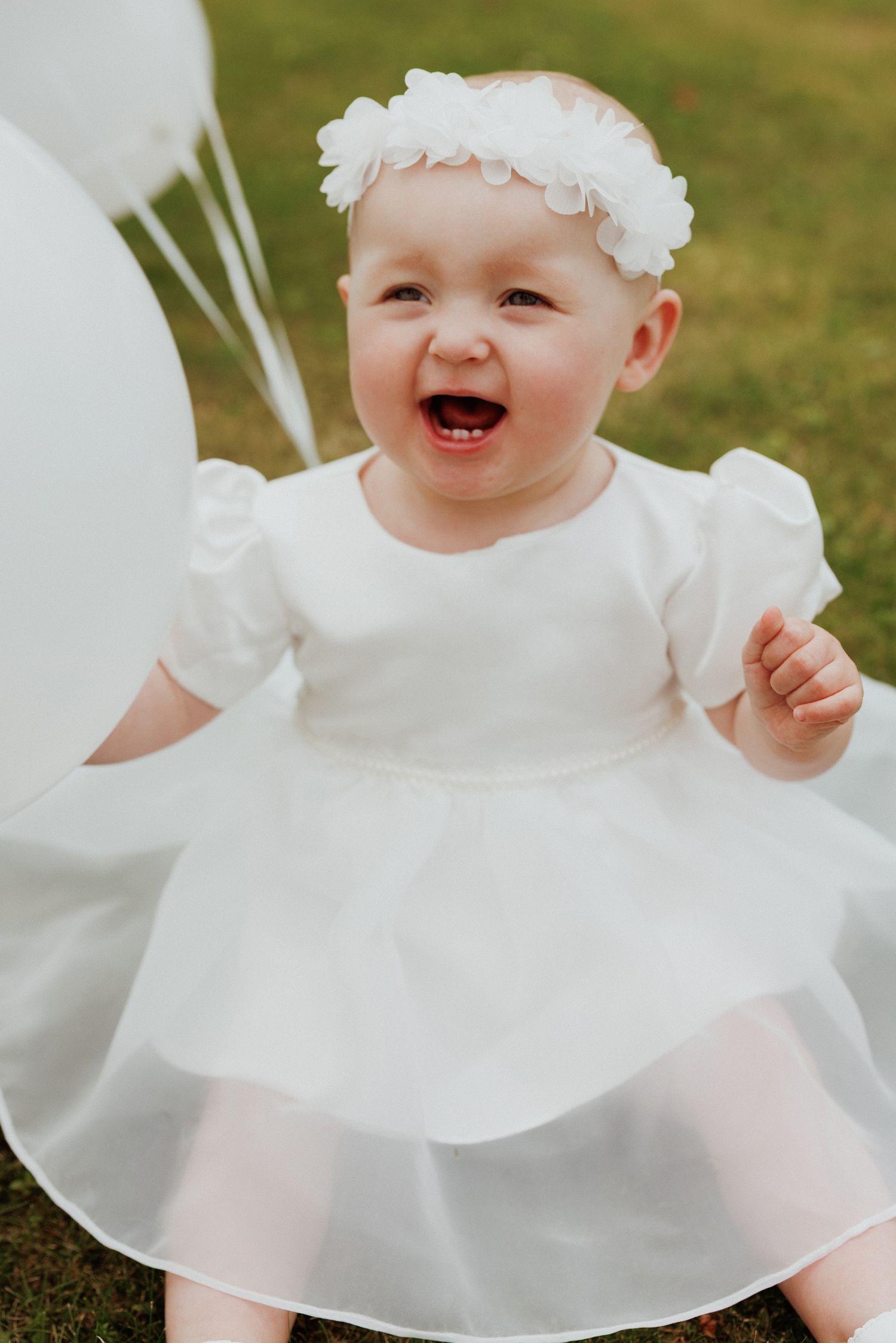 Joyful baby wearing white christening dress and floral headband smiles while playing with balloons outdoors.