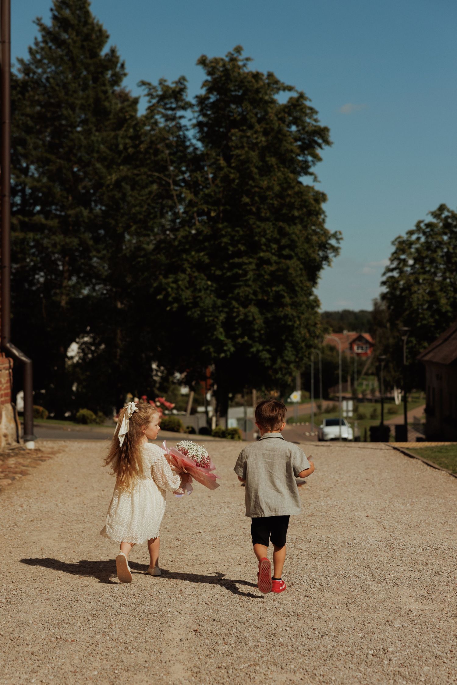 Children in white dresses playing together on a gravel path with trees in the background.