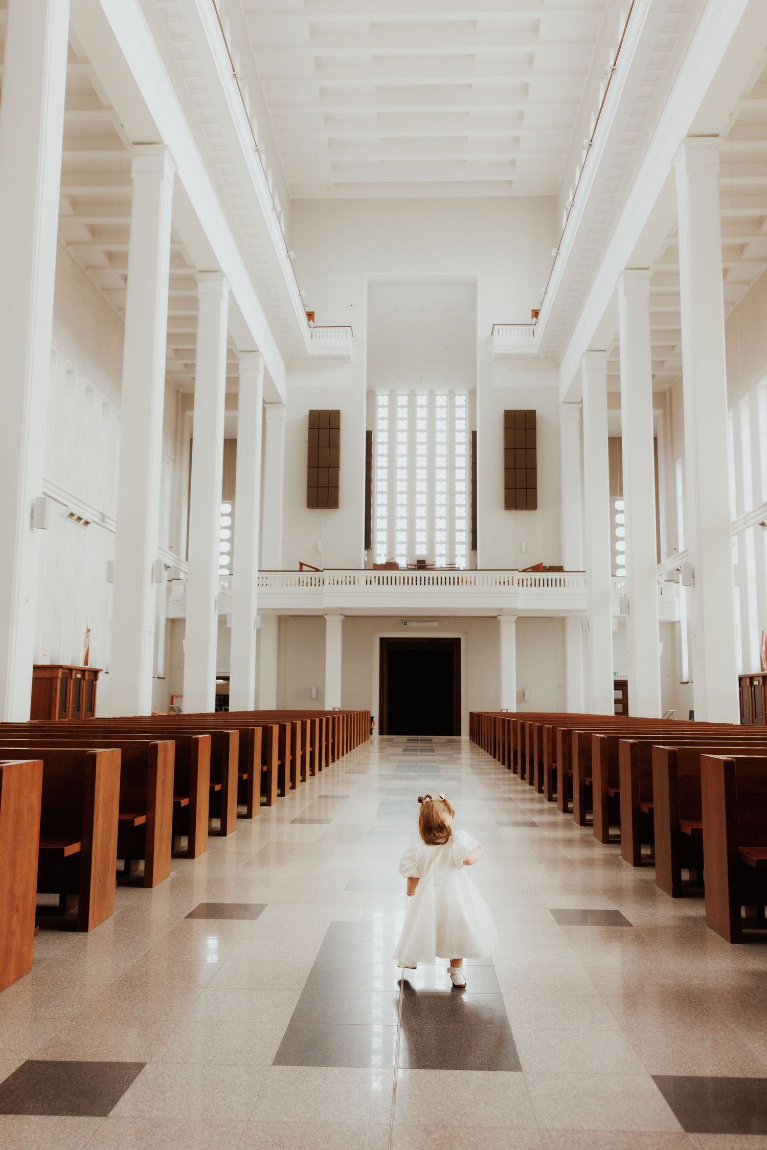 Small child in white dress walking down empty church aisle between wooden pews beneath tall white columns and glass block wall.