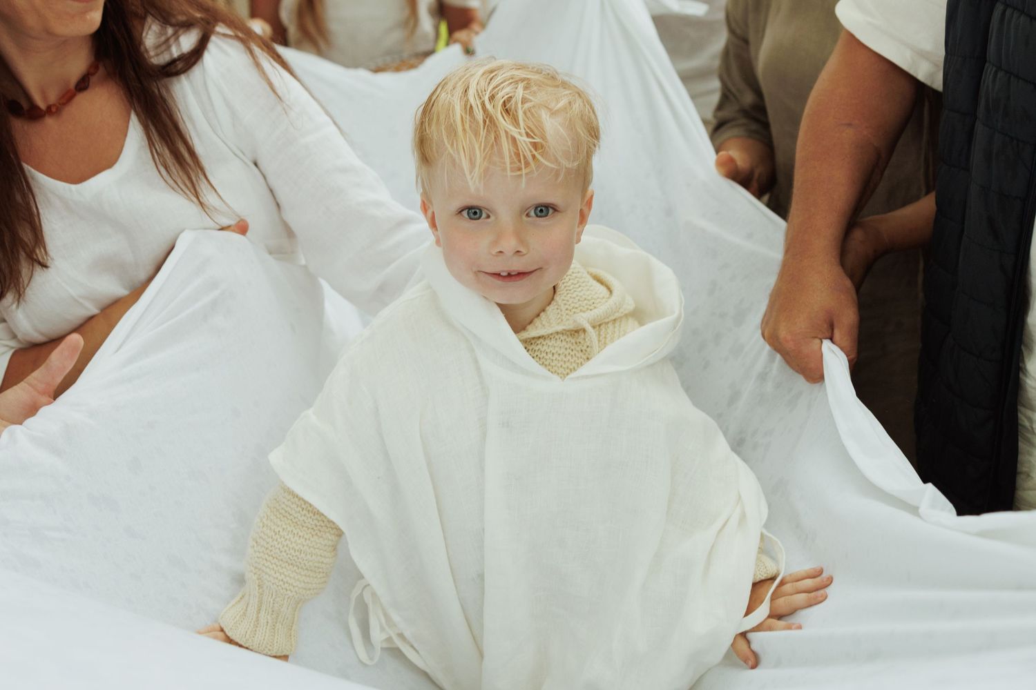 Smiling toddler wrapped in a white hooded robe surrounded by adults in white clothing.