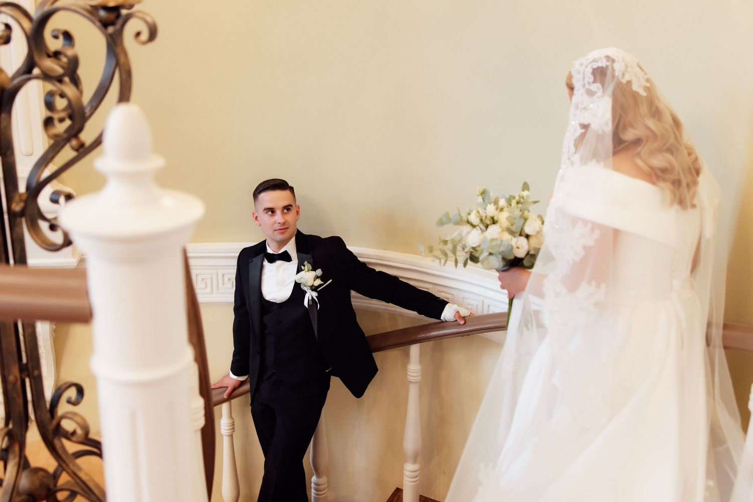 Groom in black tuxedo gazes at bride in white wedding gown and veil descending elegant staircase.