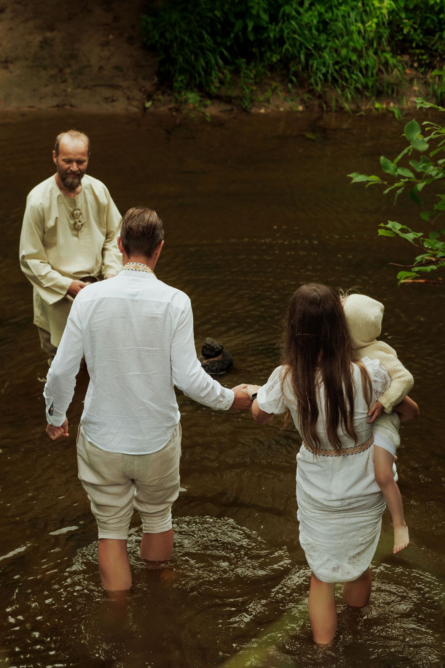 Family in white traditional clothing wading through a shallow river during a baptism ceremony.