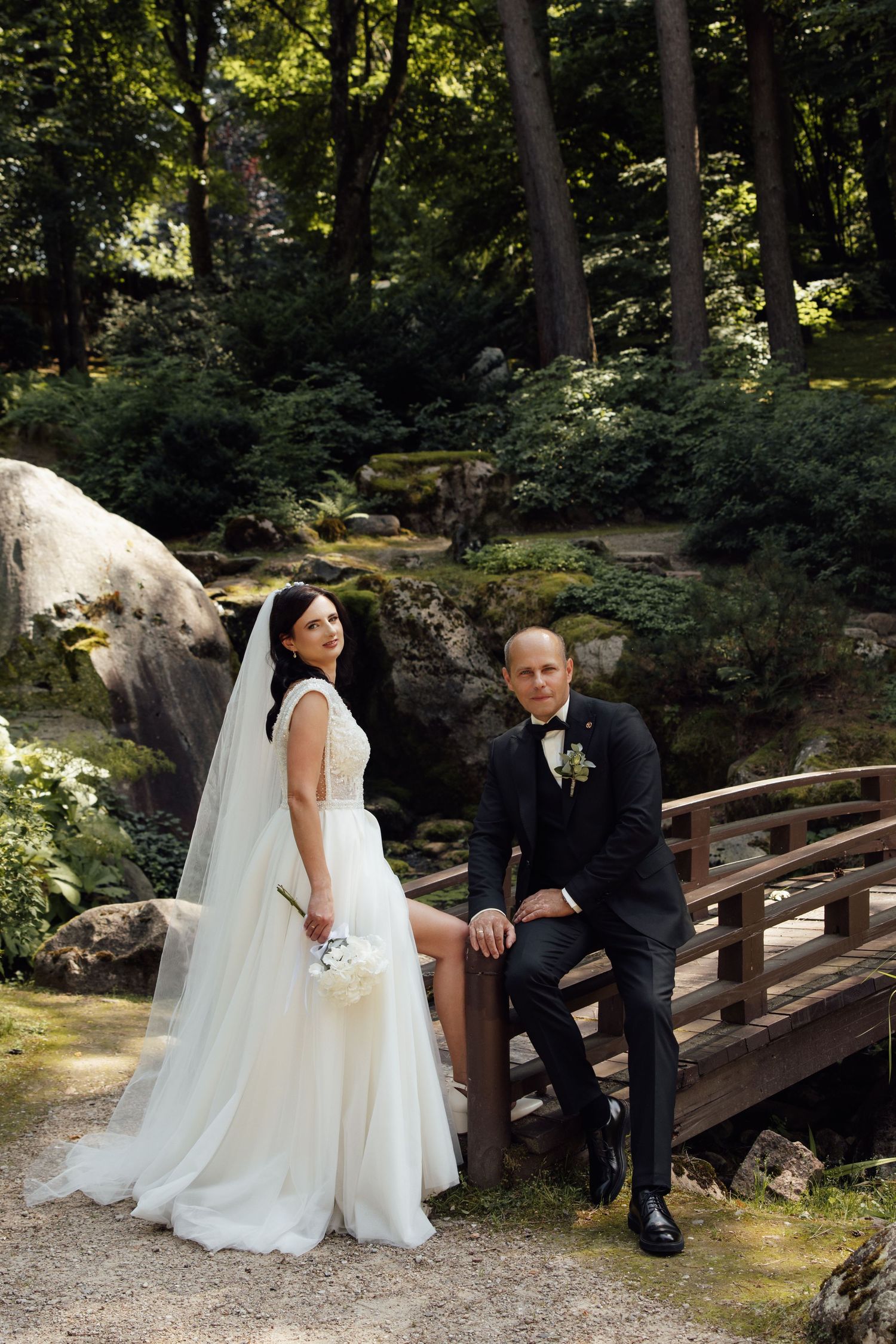 Bride in white dress and groom in black suit posing on wooden bridge in scenic forest garden.