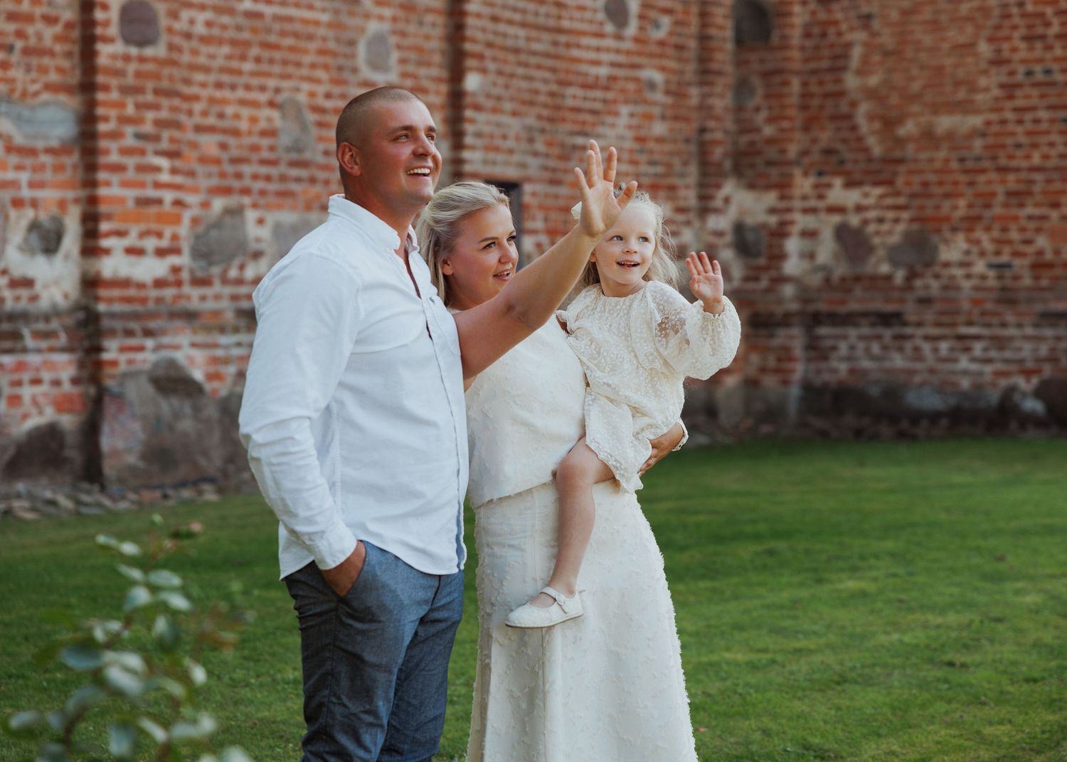 Family wearing white outfits poses together against a rustic brick wall and green lawn.