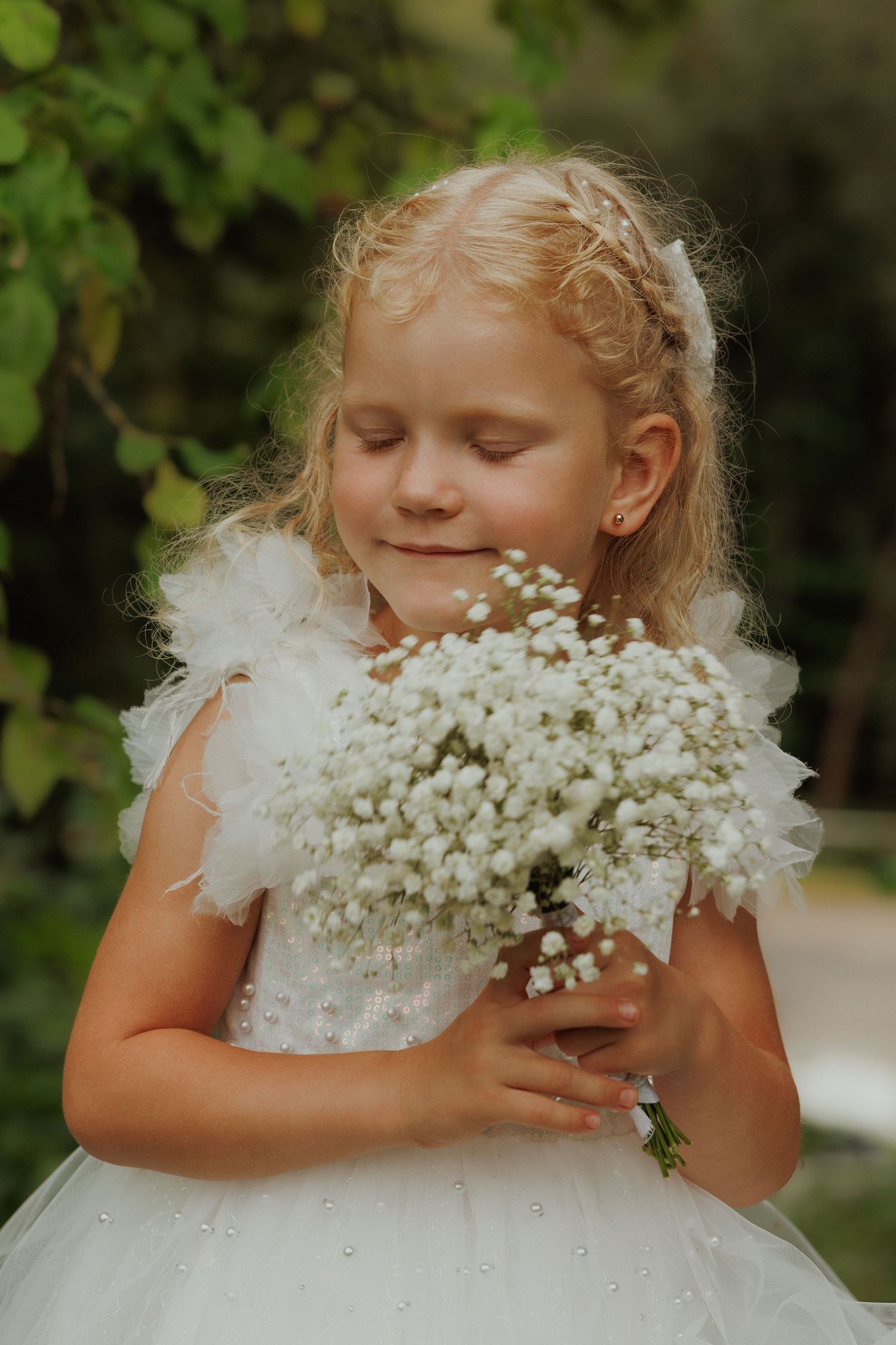 A child in a white dress buries their face in a bouquet of delicate baby's breath flowers in a garden setting.