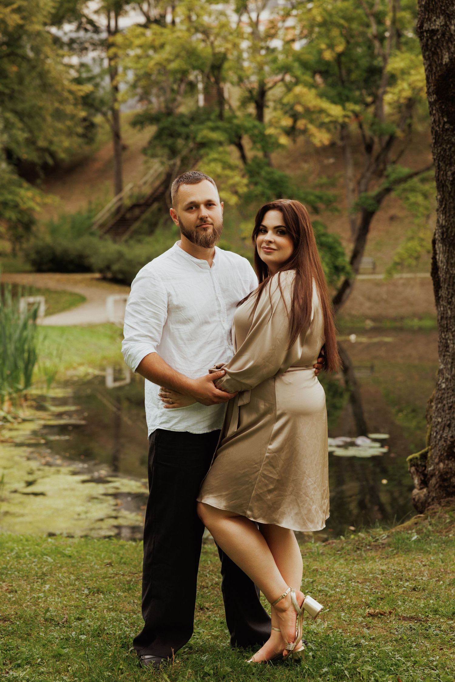 Couple poses romantically by a pond in a scenic park setting during golden hour.