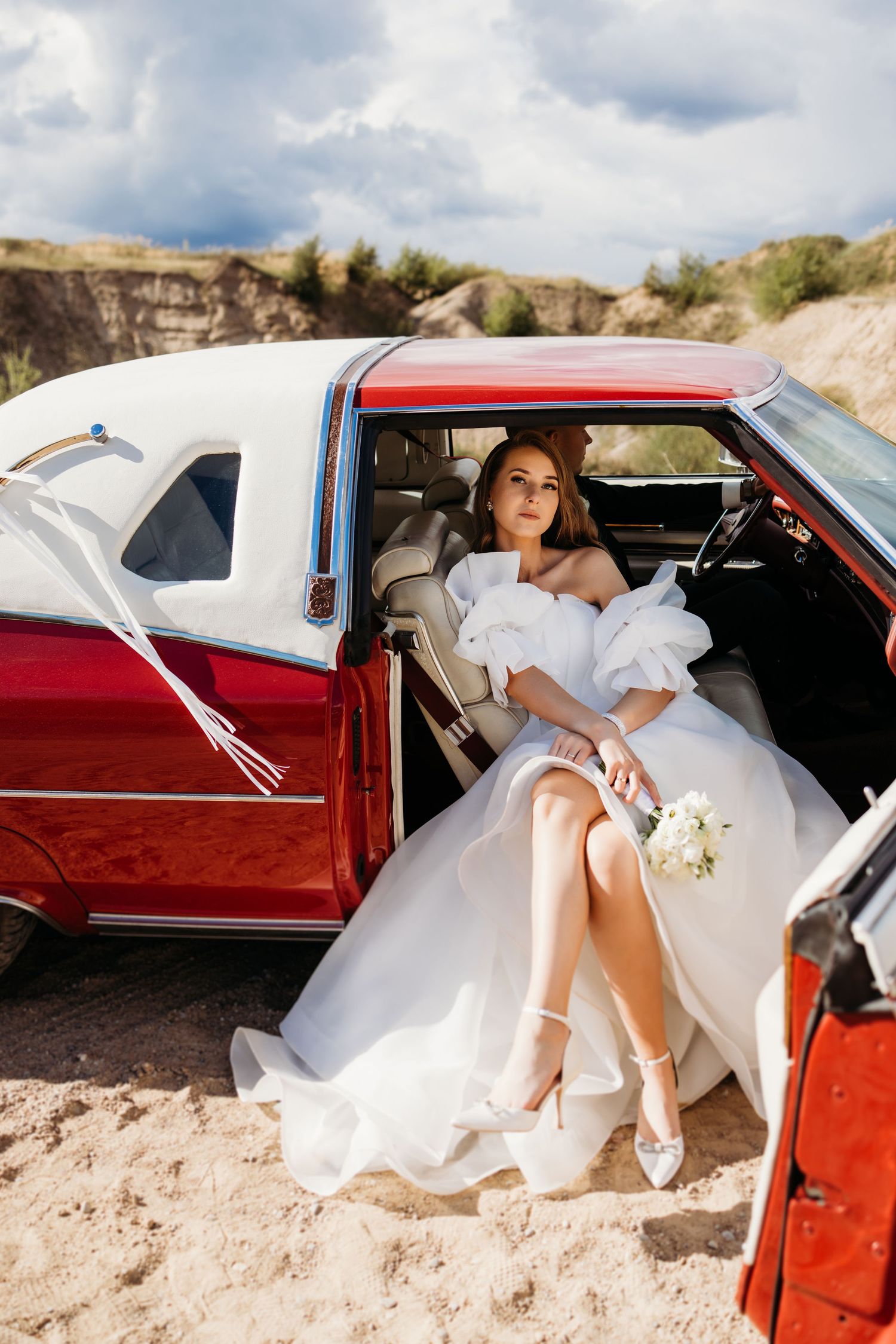 Artistic series of bride in white dress posing in vintage red car against desert backdrop.