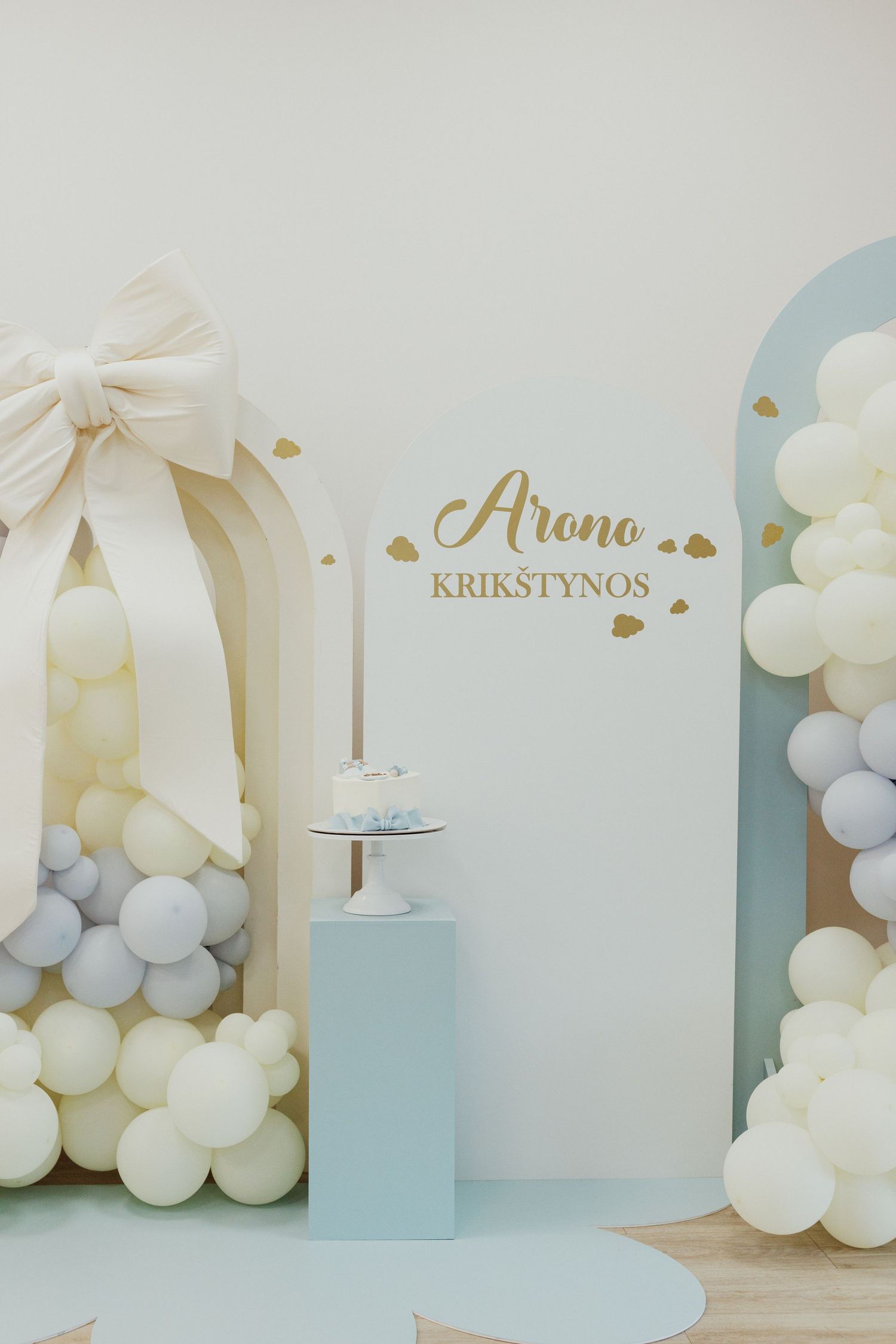 Baby shower decoration featuring white balloons, blue accents, and gold lettering against a pastel wall.