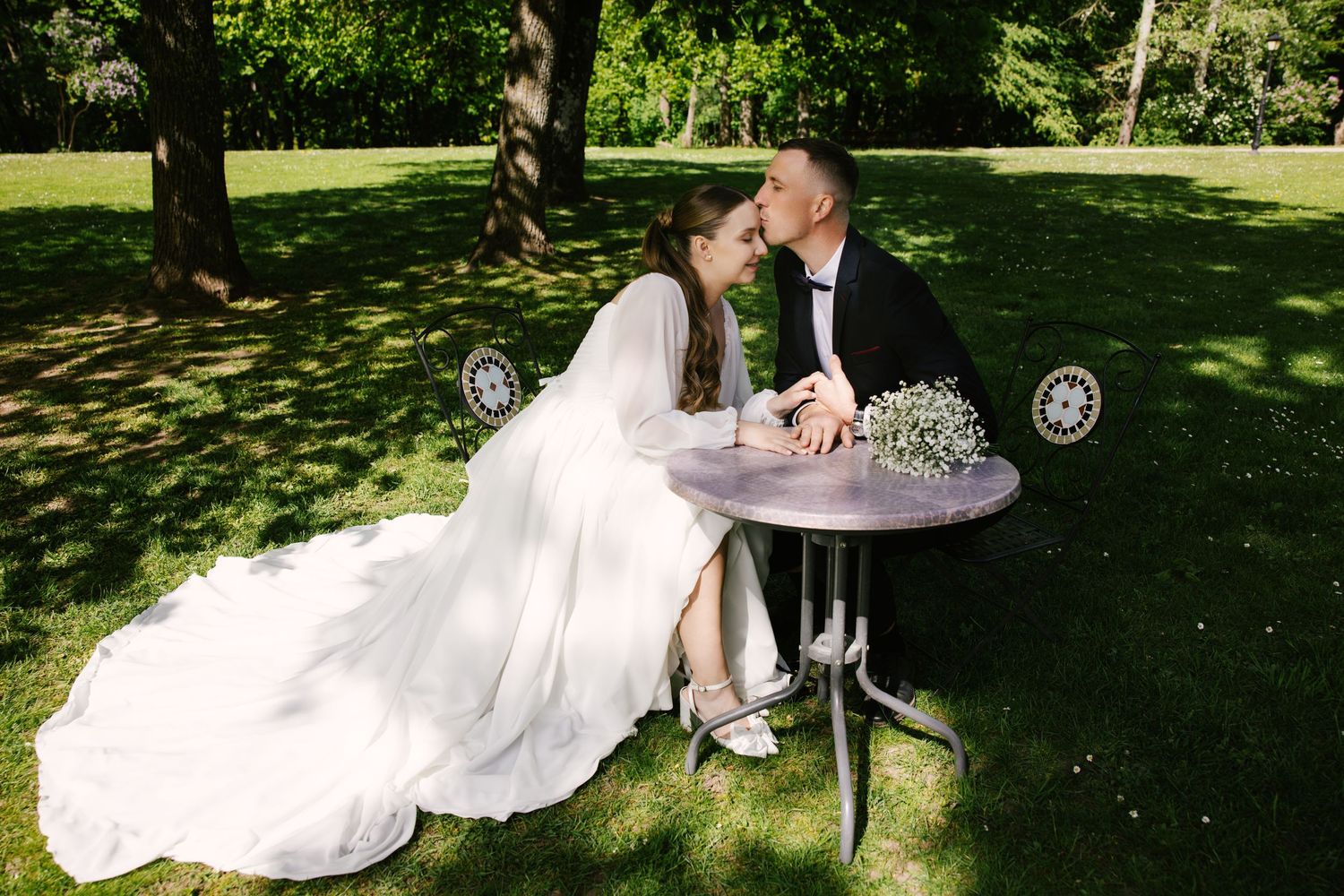 A romantic wedding moment captured at a small bistro table under shady trees in a garden setting.