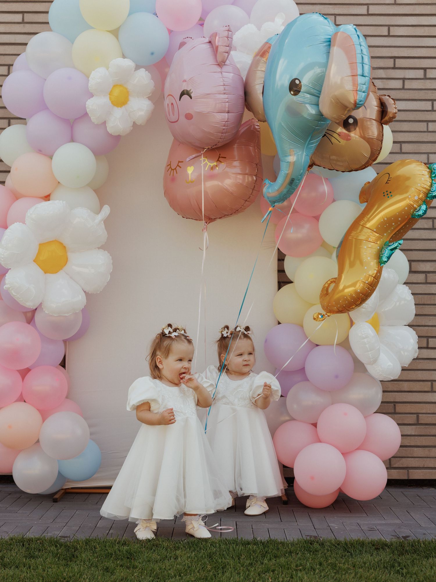 Children in white dresses pose in front of a pastel balloon arch decorated with soft pink and blue colors.