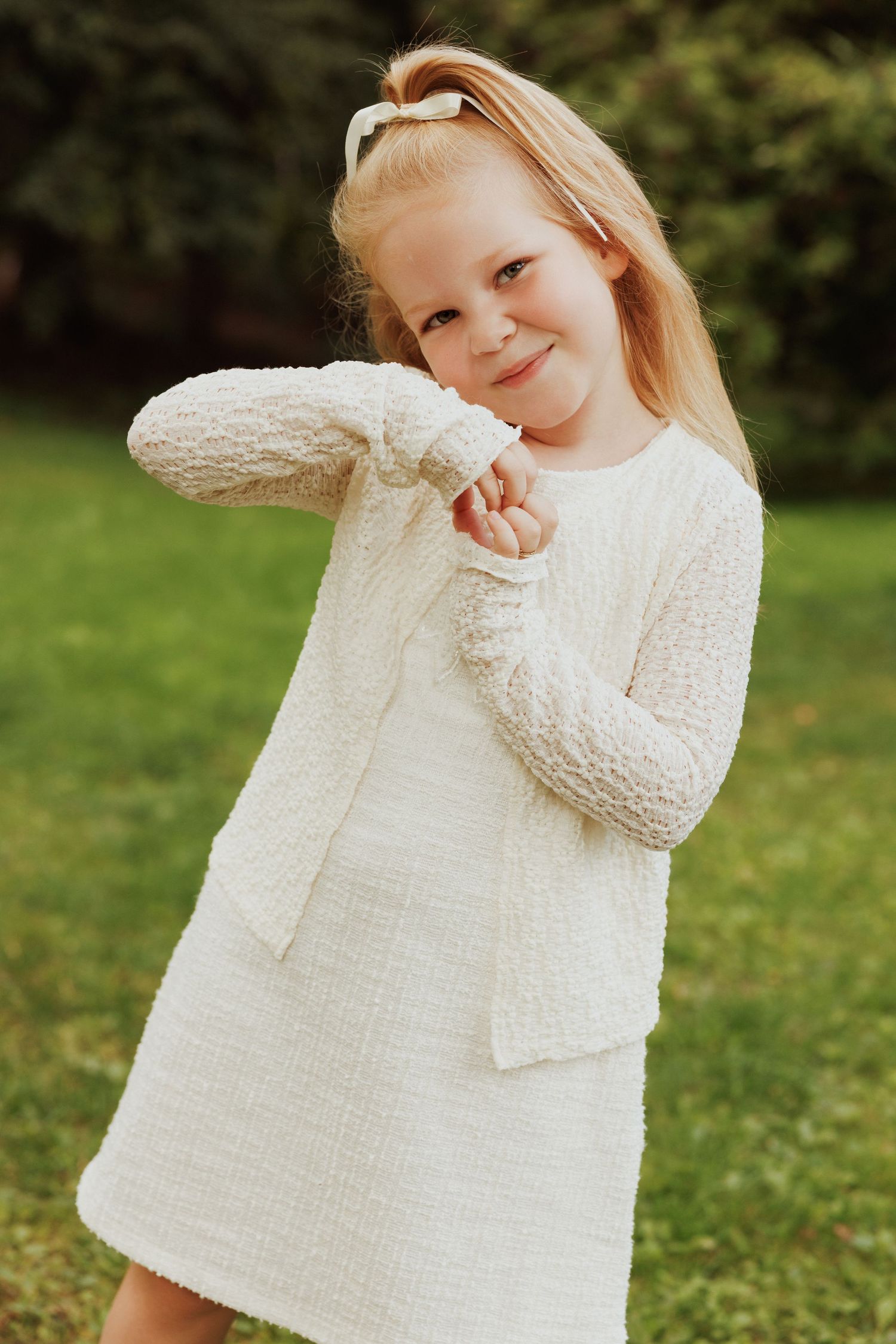Young child in white knit dress poses playfully on grass.