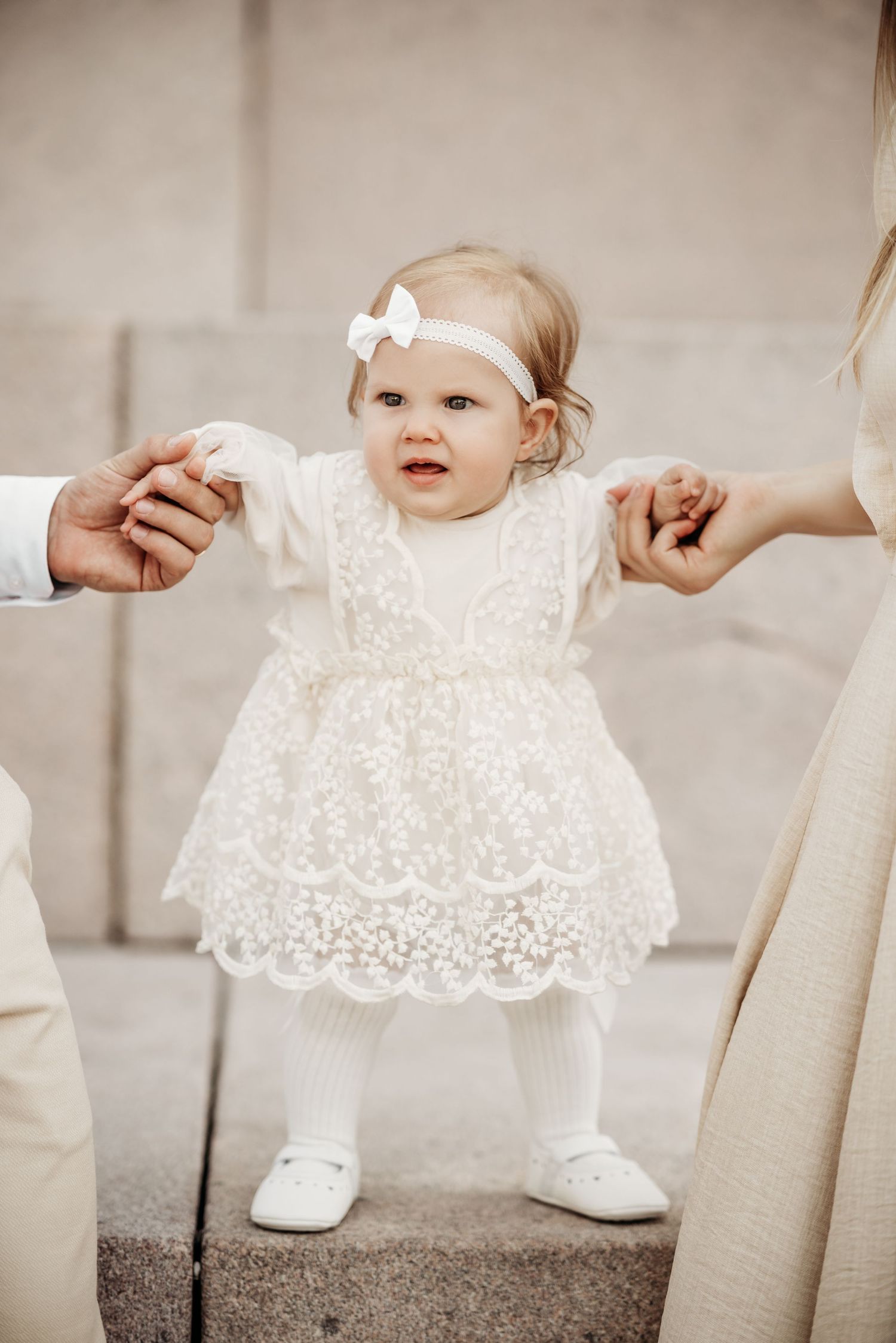 A small child in a white lace dress and tights takes first steps while holding adult hands for support.