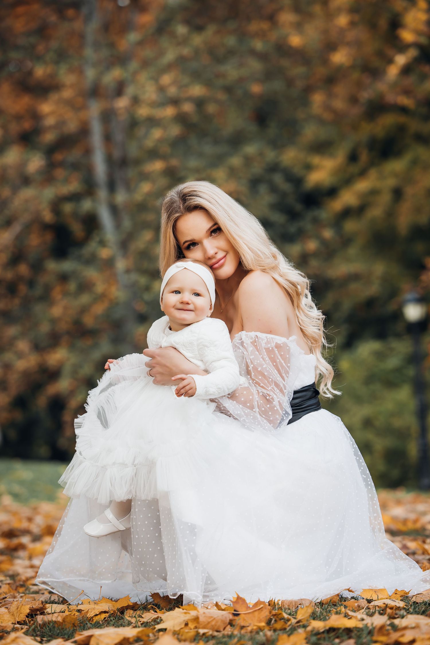 A mother and baby in matching white dresses sit among fallen autumn leaves in a park setting.