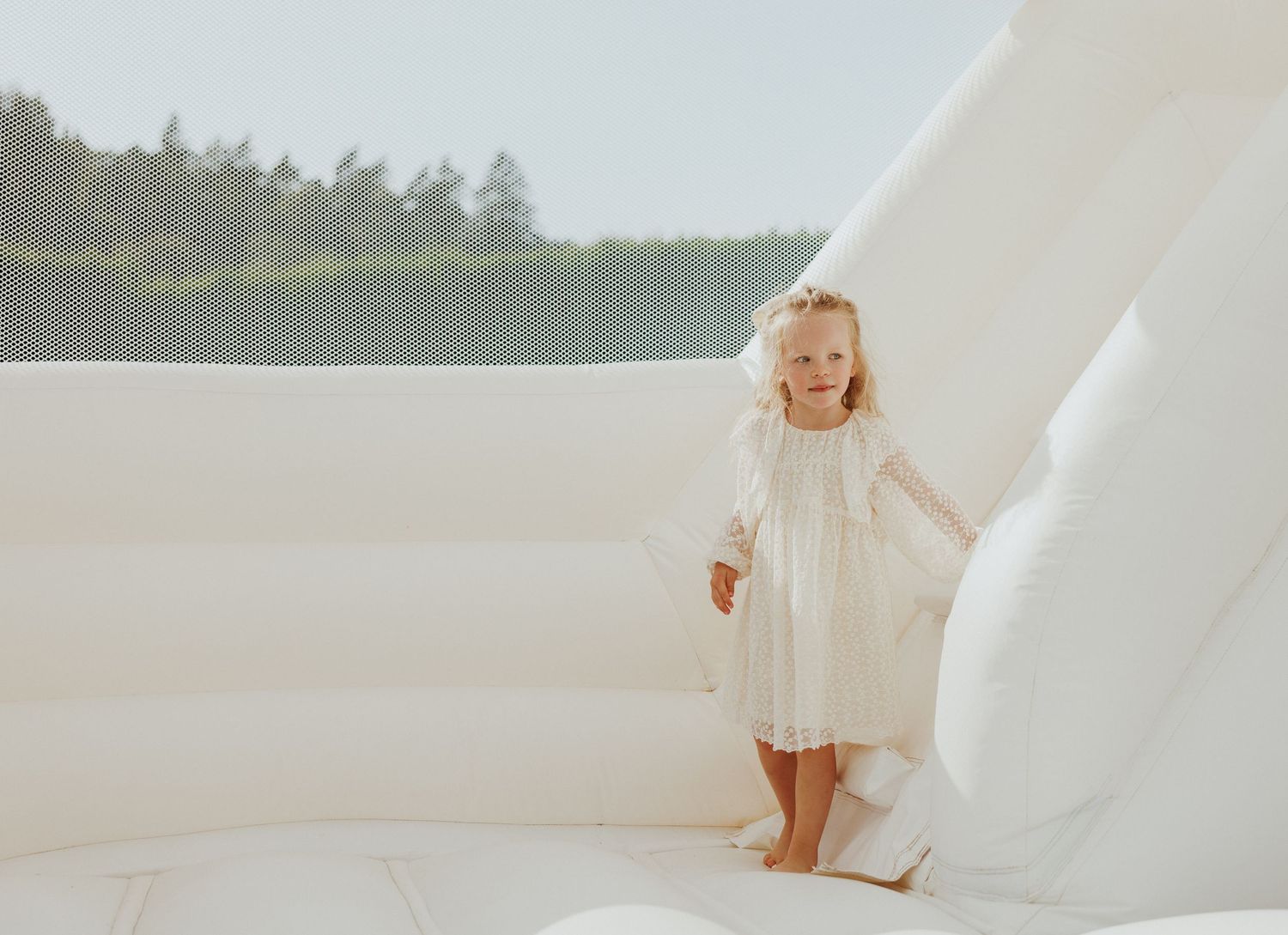 Child in white dress standing on white surface with dreamy natural backdrop.
