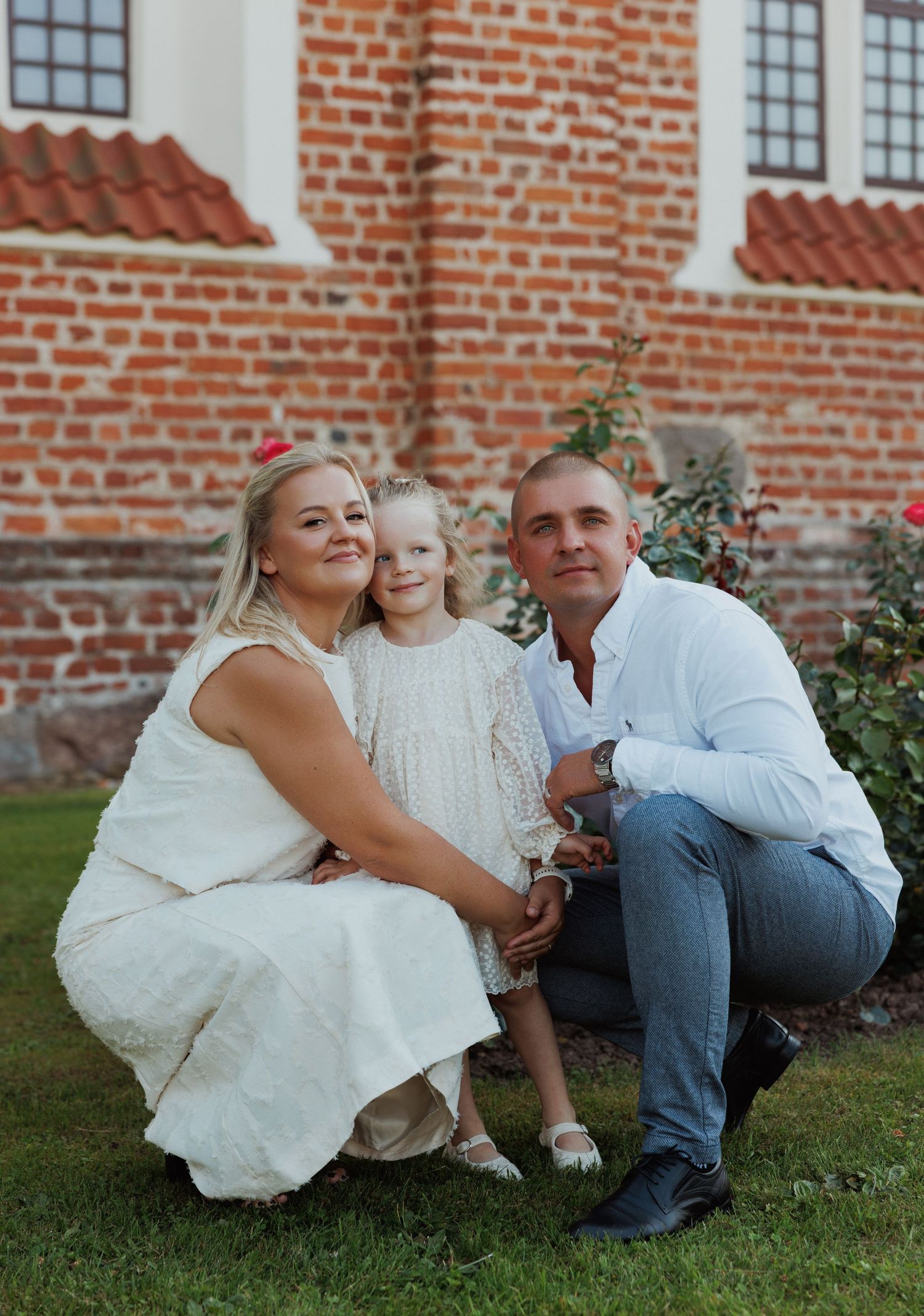 Family poses together on grass against brick wall in coordinated white and denim outfits during outdoor photo session.