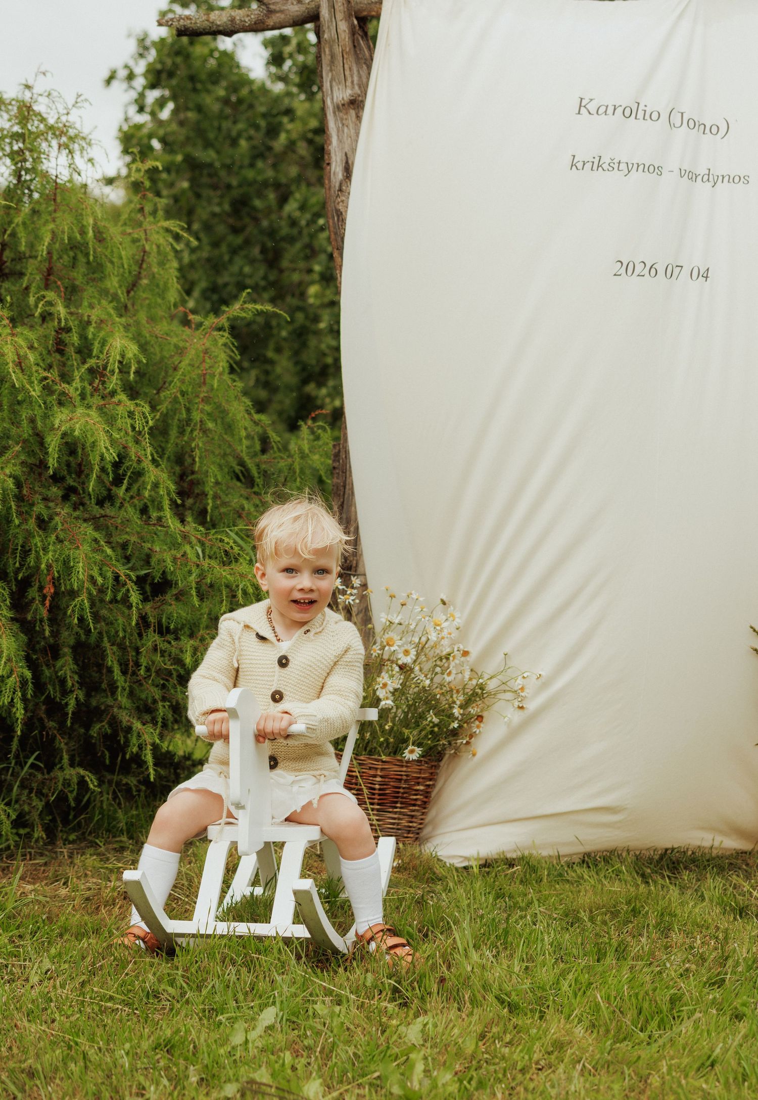 Smiling toddler boy in beige knit outfit sitting on white rocking horse in garden at baptism celebration.