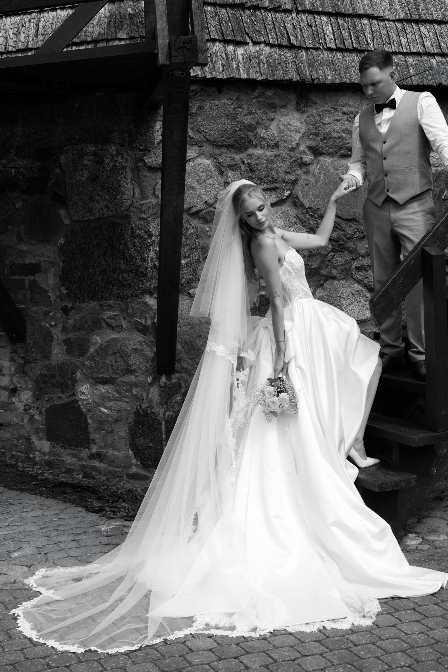 Black and white wedding photo of bride in flowing gown and veil with groom in suit on stone steps.