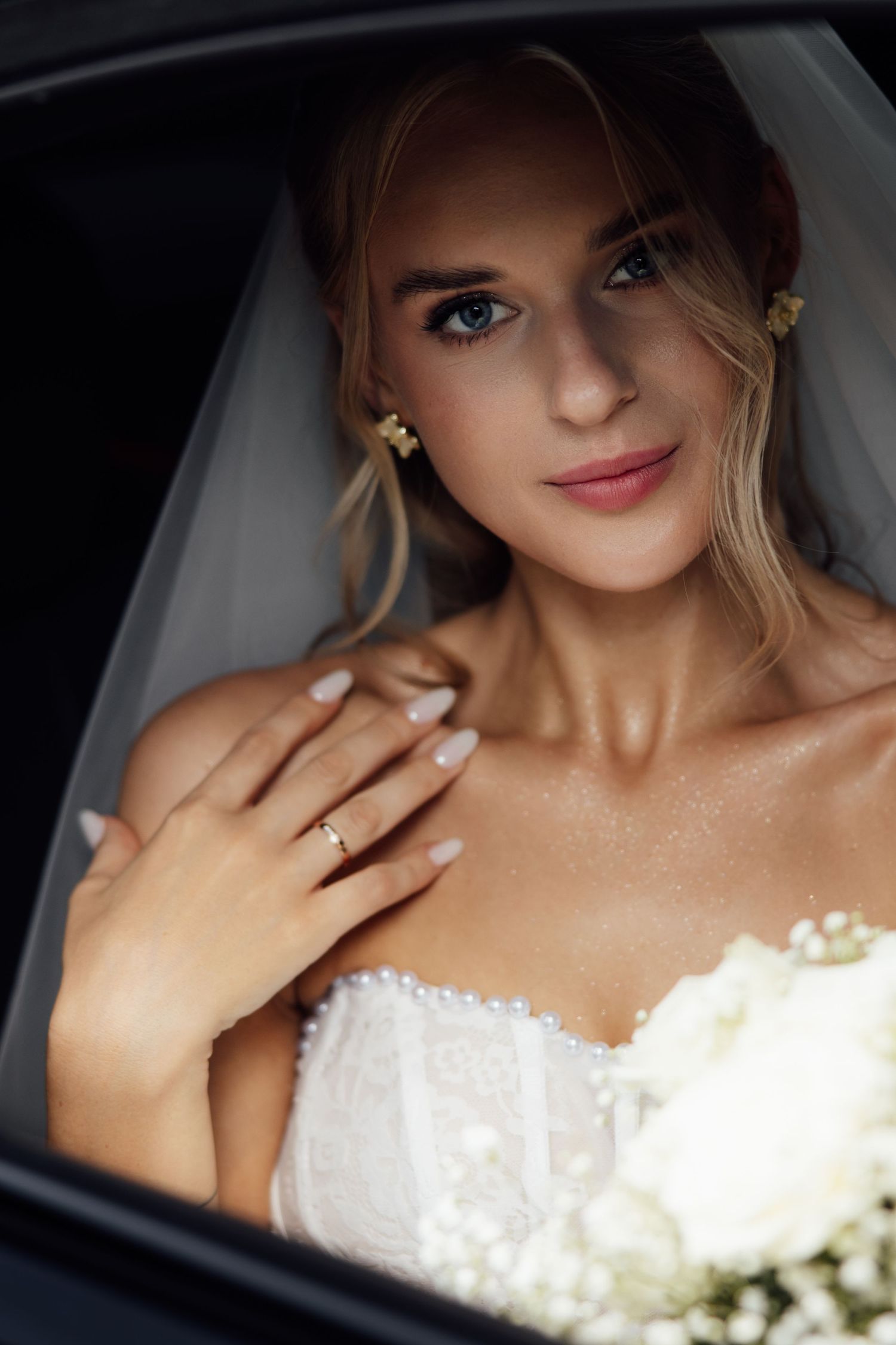 Elegant blonde bride in white strapless gown holding bouquet, wearing ring and pearl earrings, posing in car window.