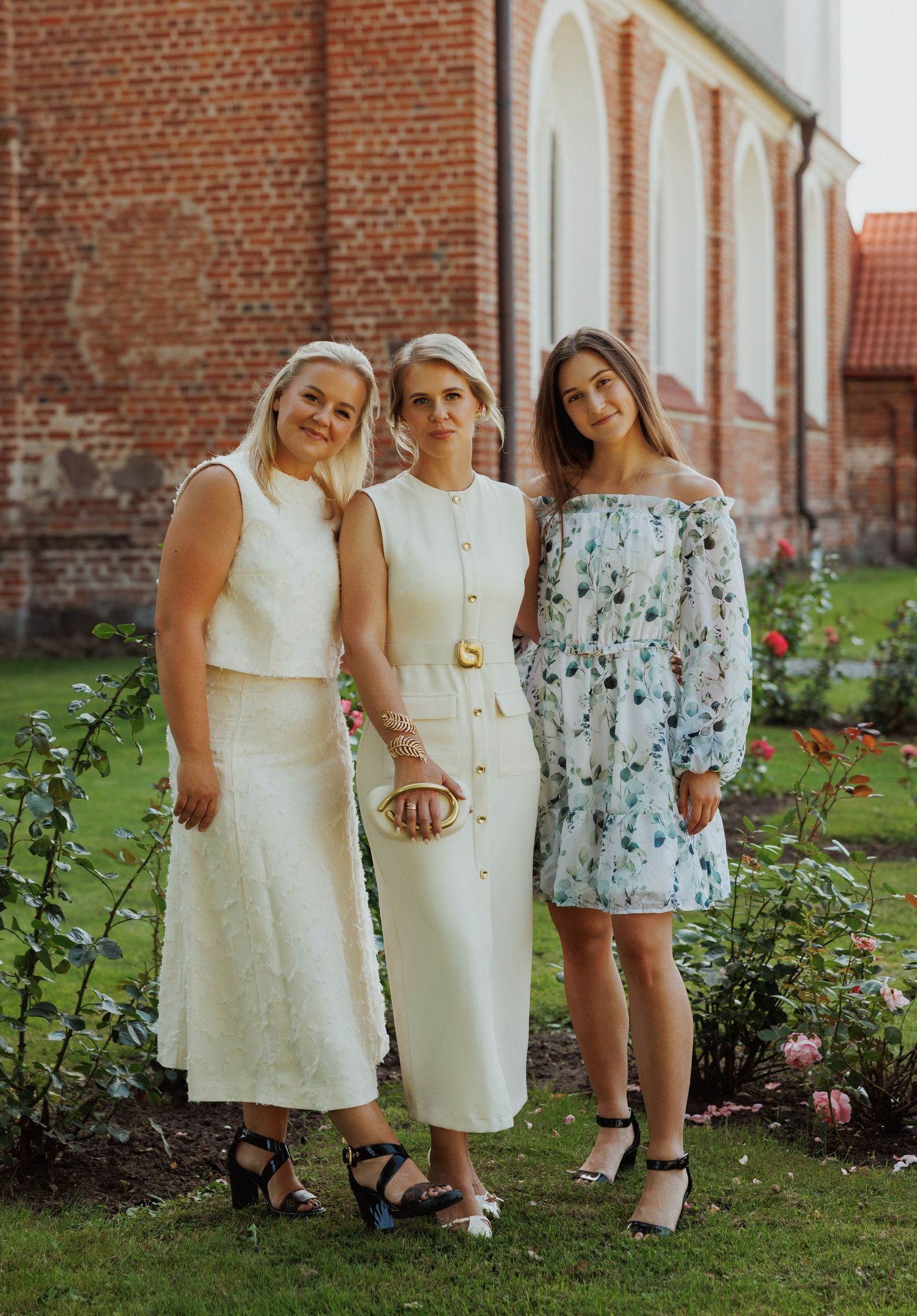 Children in white dresses and floral outfits stand together in a garden beside a brick building.