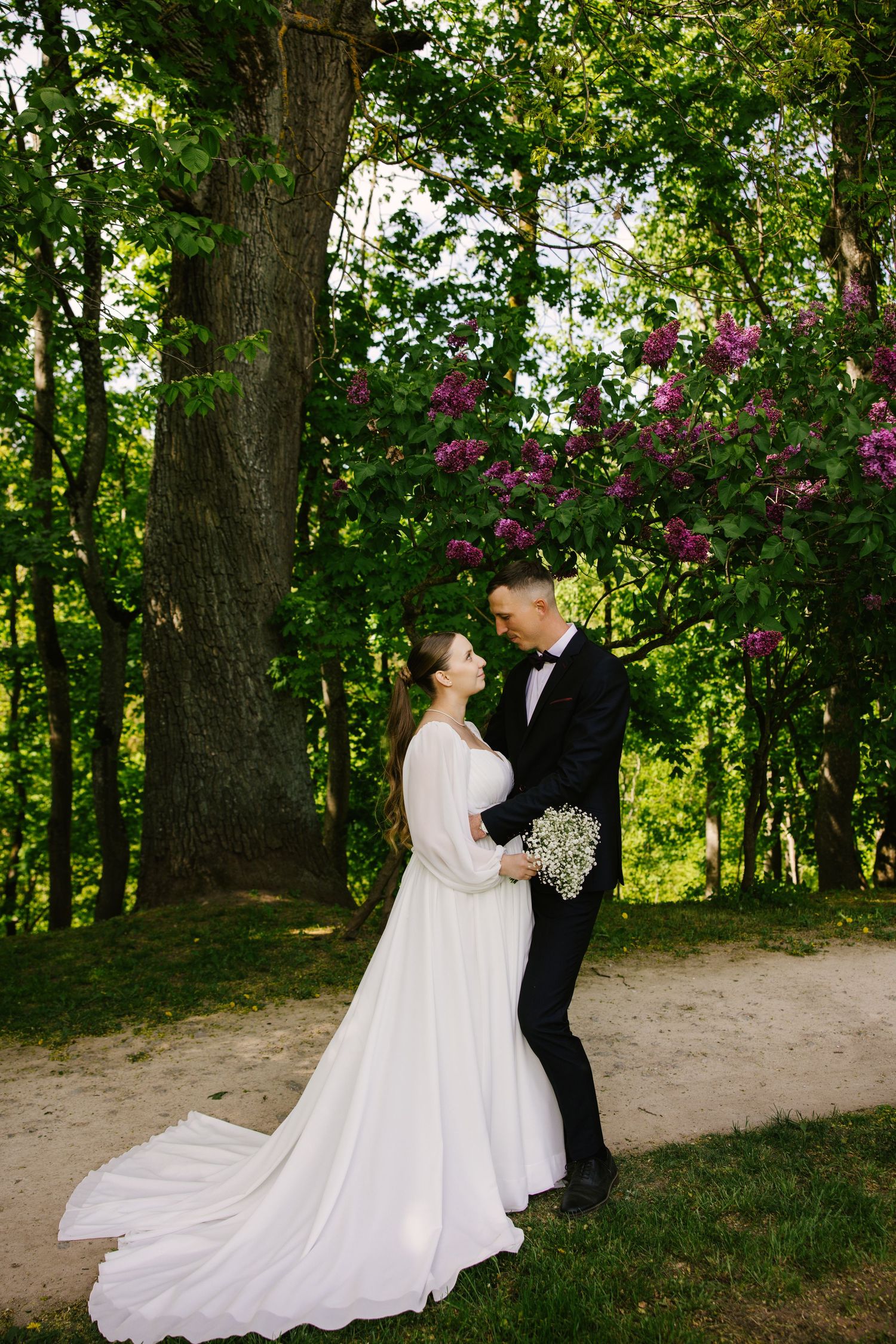 A wedding couple poses on a garden path surrounded by blooming spring trees and greenery.