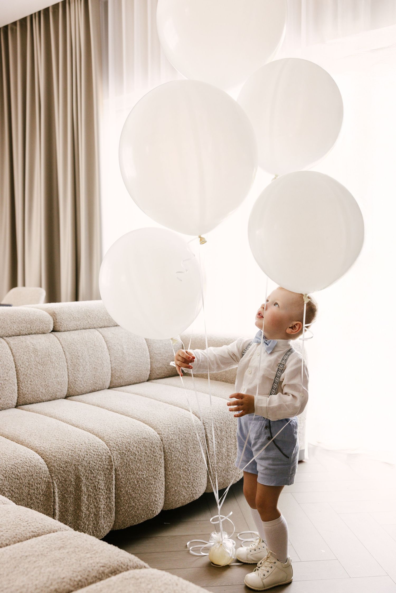 Small child plays with white balloons next to modern grey sectional sofa.