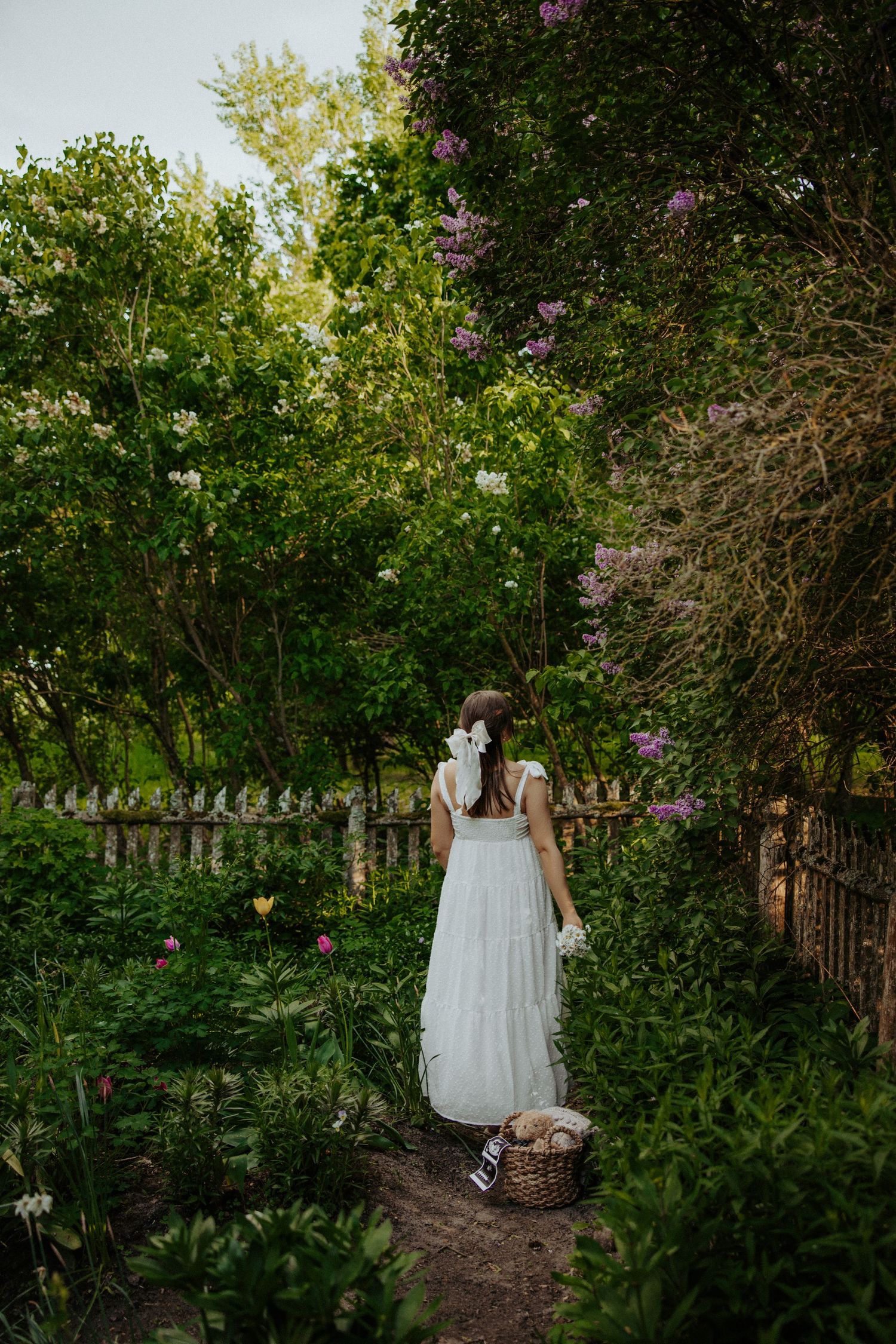 White dress figure stands in lush garden with wooden fence and blooming flowers.
