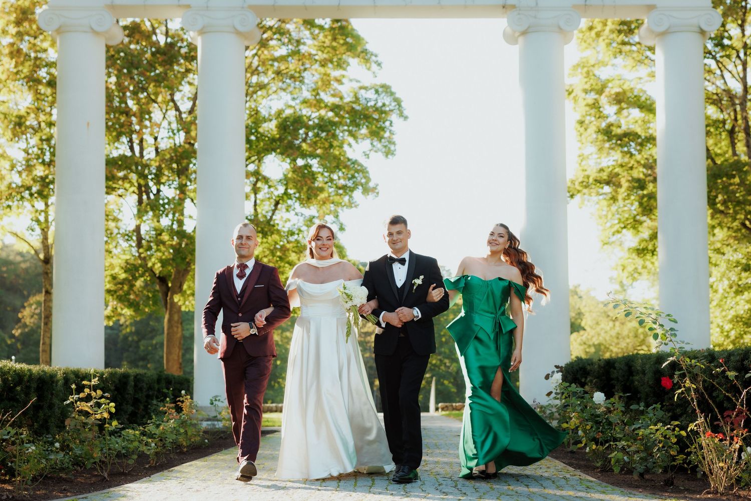 Wedding party walks along columned pathway with bride in white and bridesmaids in green.