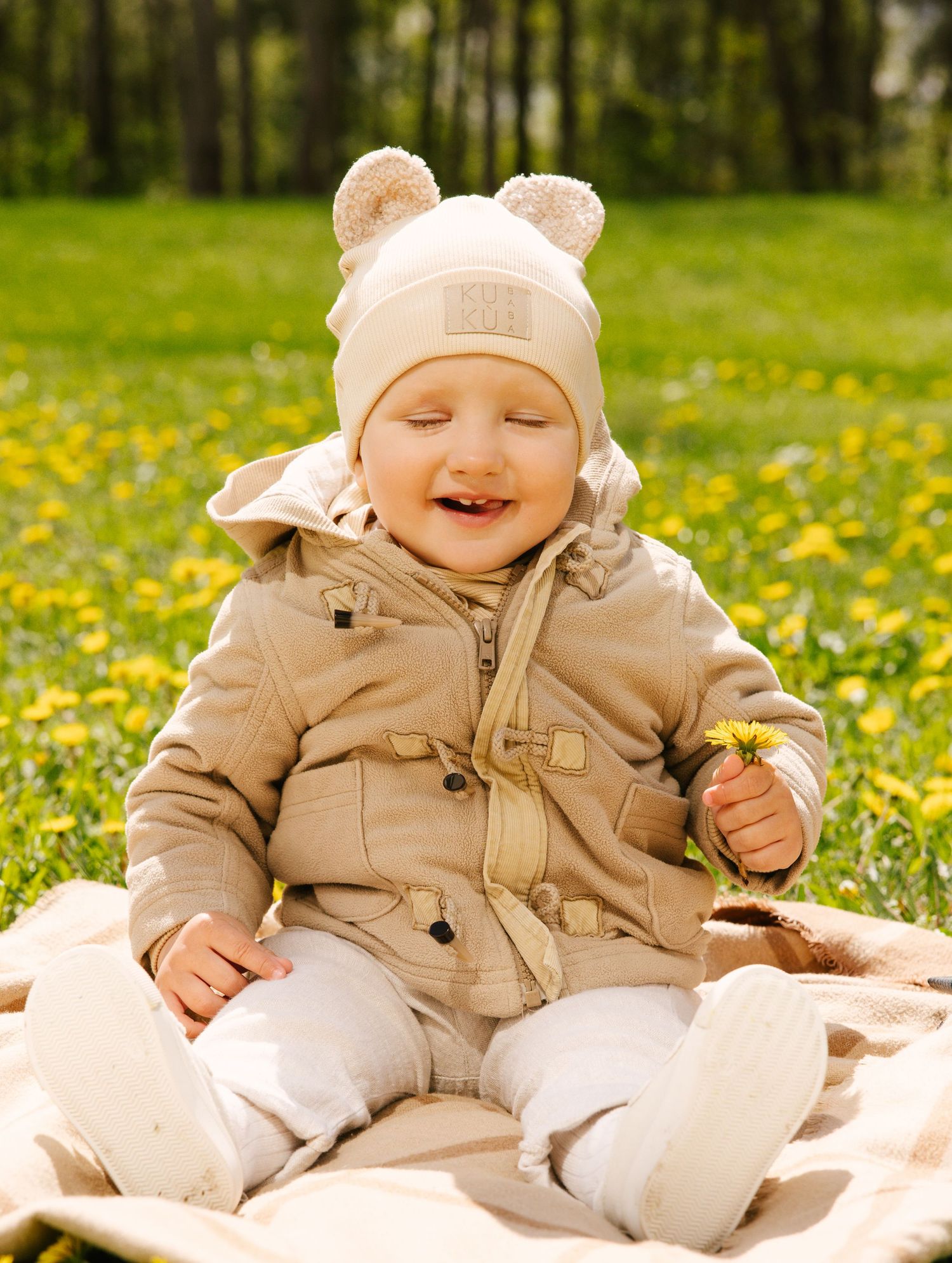 Baby wearing beige outfit and hat sits on white blanket in field of yellow dandelions on sunny spring day.